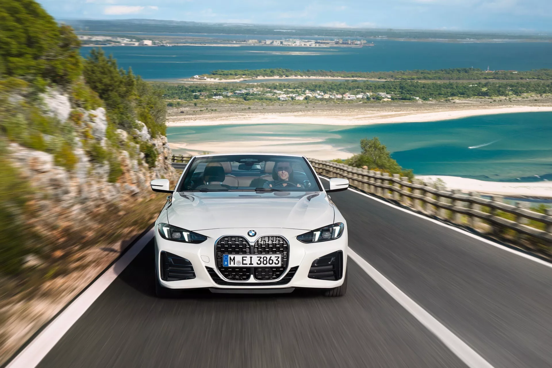 Luksuzna limuzina BMW serije 4 (G22-G23-G26-G82-G83 prenova) iz leta 2024 vozi po slikoviti obalni cesti, ki prikazuje njeno veliko ledvično rešetko, kotne LED žaromete in dinamično oblikovanje sprednjega odbijača