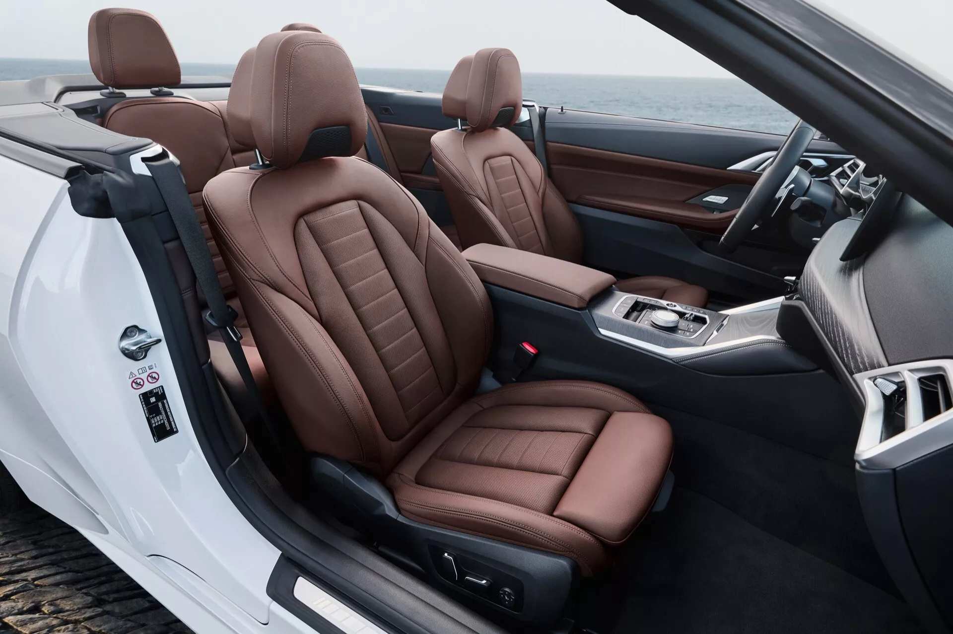 Notranjost BMW serije 4 2024 (G22-G23-G26-G82-G83 facelift) prikazuje sprednjo kabino iz zadnje-stranske perspektive, poudarjajoč razkošne rjave usnjene sedeže, kabriolet naravo in napredne kontrole na vratih in na osrednji konzoli.
