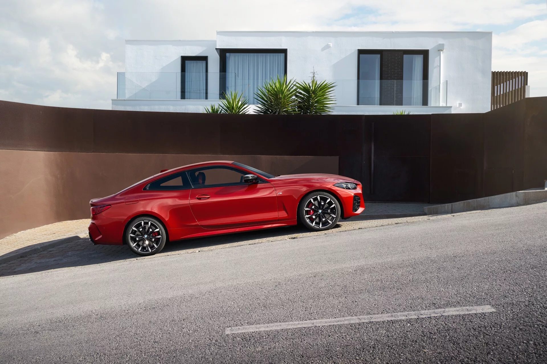 Svetlo rdeč dvovratni kupe BMW serije 4 (G22-G23-G26-G82-G83 Facelift) letnik 2024 parkiran na robu ulice, ki prikazuje svoj elegantni, športni dizajn z veliko izrazito rešetko, LED žarometi, večkraka zlitinska kolesa, nizkoprofilne pnevmatike, visokokakovostni sijoč lak, bočna ogledala z integriranimi smerniki in visokozmogljiv zavorni sistem v stanovanjskem okrožju.