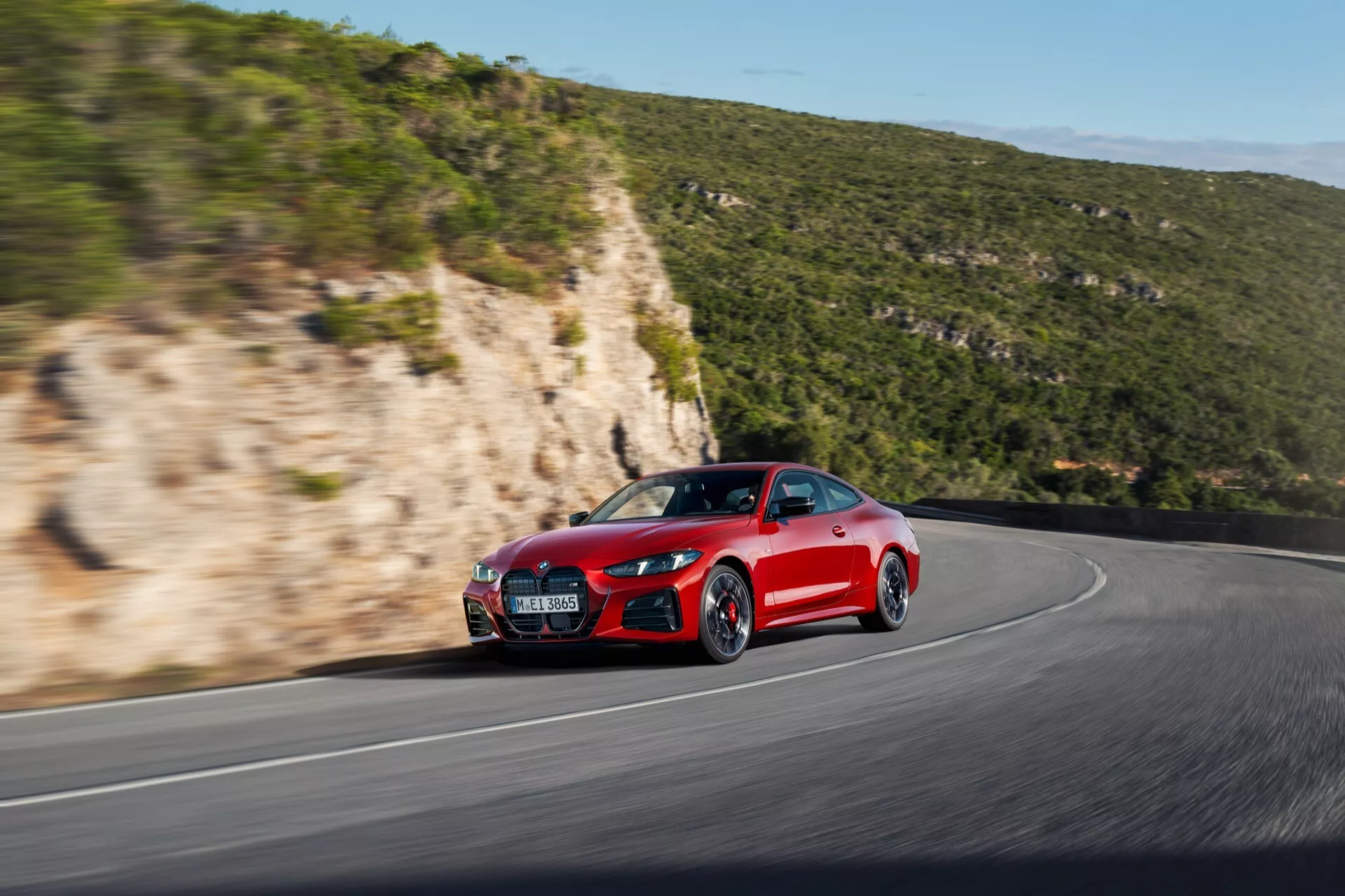 Dinamični bočni sprednji pogled na rdeči športni avto BMW serije 4 2024 (G22-G23-G26-G82-G83 prenova), ki prikazuje njegov agresivni slog, aerodinamične linije, športni sprednji odbijač, pokrov motorja, stranski panel, sprednje kolo in pnevmatiko na ovinkasti cesti