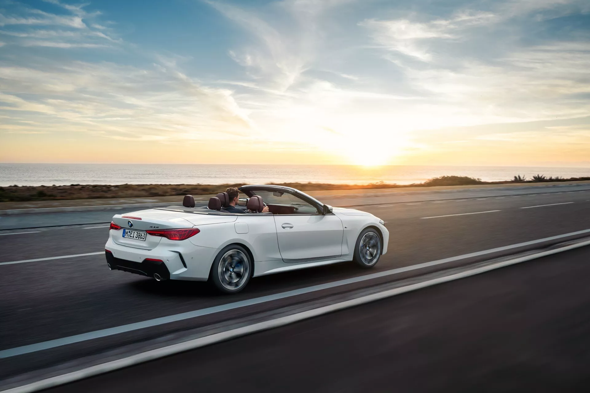 BMW serije 4 kabriolet (preoblikovanje G22-G23-G26-G82-G83) v letu 2024 na obalni cesti ob sončnem zahodu, zračni pogled od zadaj in zgoraj, ki prikazuje njegovo belo zunanjost, zloženo streho kabrioleta in športno zasnovo
