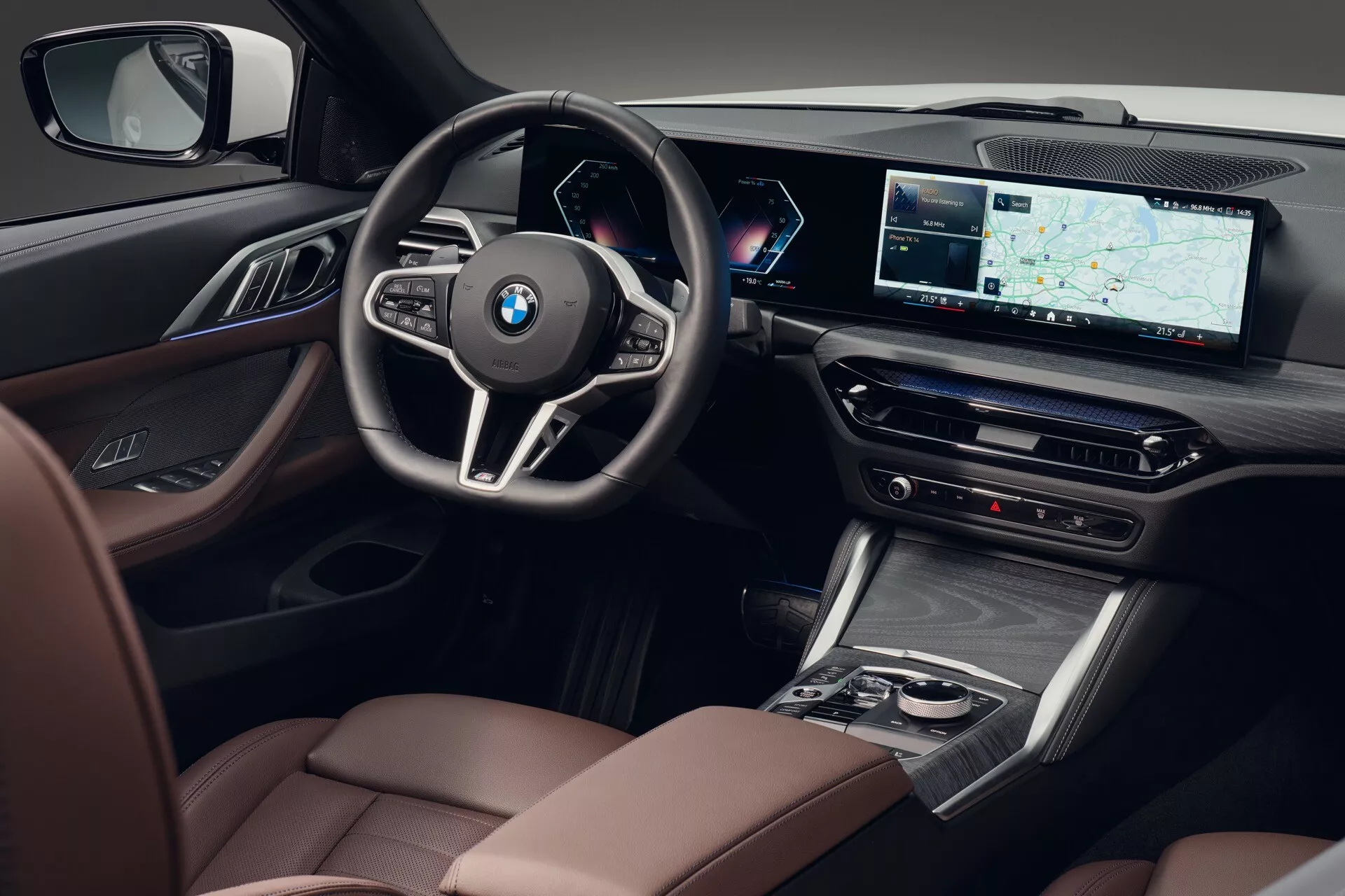 Notranji posnetek z rahlo povišanega kota, usmerjen proti voznikovi strani BMW serije 4 2024 (G22-G23-G26-G82-G83 prenova), ki prikazuje sedeže z visokokakovostnim usnjem z nasprotnimi šivi, leseni ali leseni učinek obrobe, kontrolnike premium zvočnega sistema na vratni plošči, športni volan s tremi kraki z multifunkcijskimi kontrolniki in značko BMW, povsem digitalni in prilagodljivi instrumentni zaslon za volanom, velik širokozaslonski infotainment zaslon z GPS navigacijo, kontrole za klimatizacijo, elegantno prestavno ročico, sredinsko konzolo s sledilno ploščico ali krmilnikom obdano s kromiranimi detajli in delno vidnim luksuznim sedežem sovoznika, kar poudarja tehnološke in luksuzne lastnosti.
