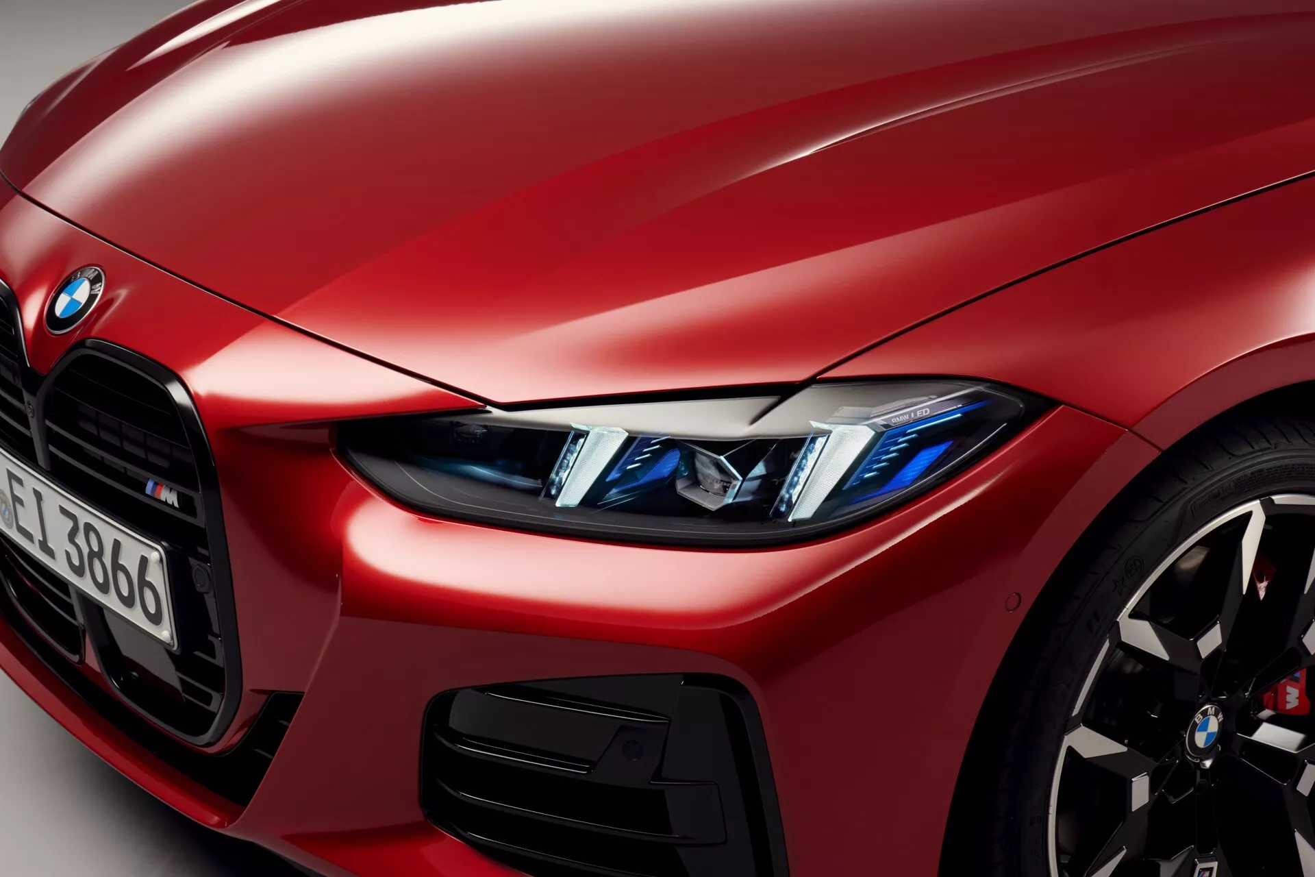Posnetek iz bližine rdečega BMW serije 4 letnik 2024 (preoblikovanje G22-G23-G26-G82-G83), ki prikazuje levi sprednji kvart avtomobila z poudarkom na LED žarometih z angelčki, delni pogled na ikonično ledvično rešetko, športno lito kolo z visokozmogljivim zavornim sistemom, metalno rdečo baroserijo in detajl zračnega dovoda ter delno vidno značko BMW in identifikacijo serije.