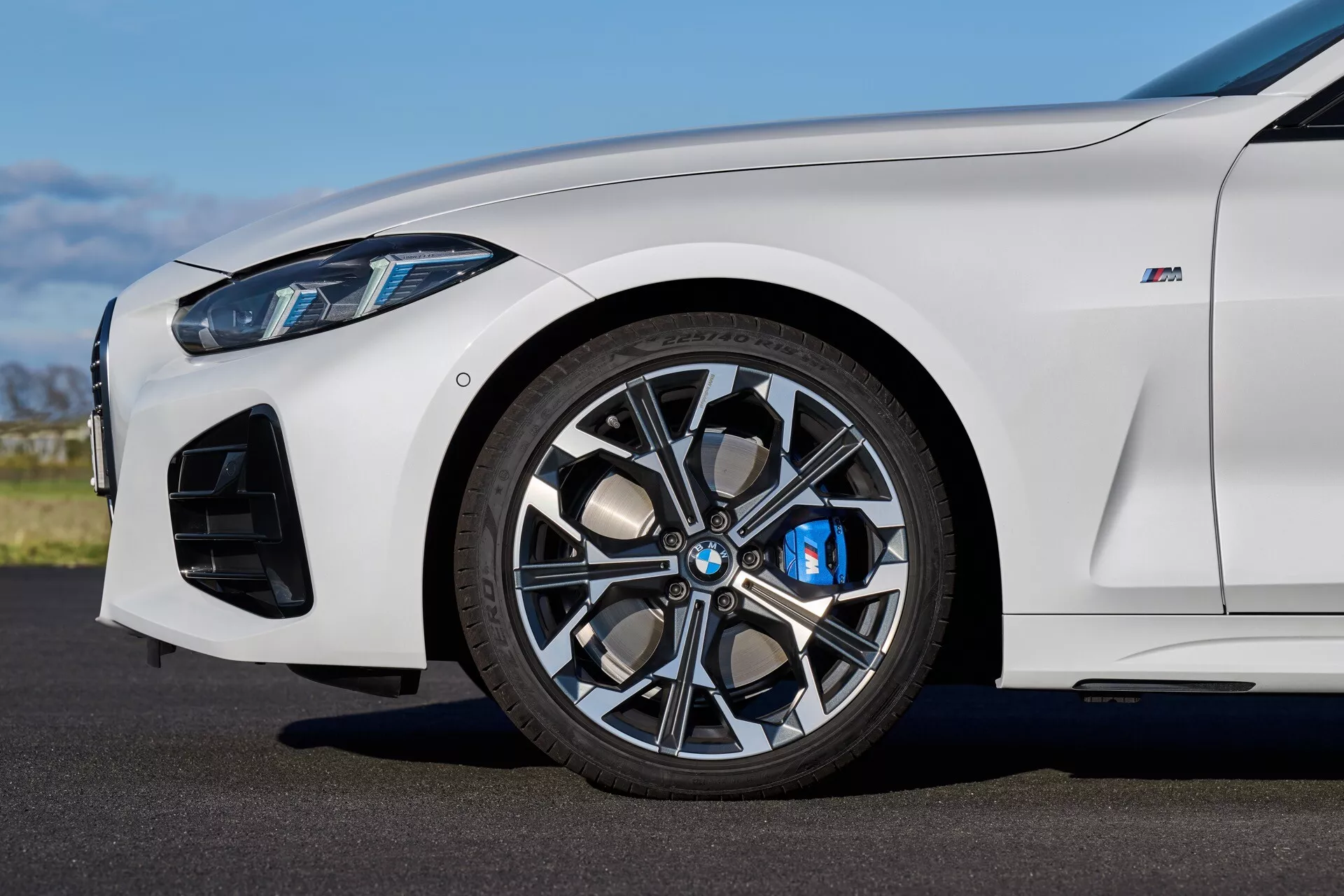 Podroben bočni pogled na beli BMW serije 4 (G22-G23-G26-G82-G83 Facelift) iz leta 2024, osredotočen na sprednje kolo z večkrakim zlitinskim obročem, modro zavorno čeljust in na zmogljivost usmerjene zračne zajeme pod naravno dnevno svetlobo