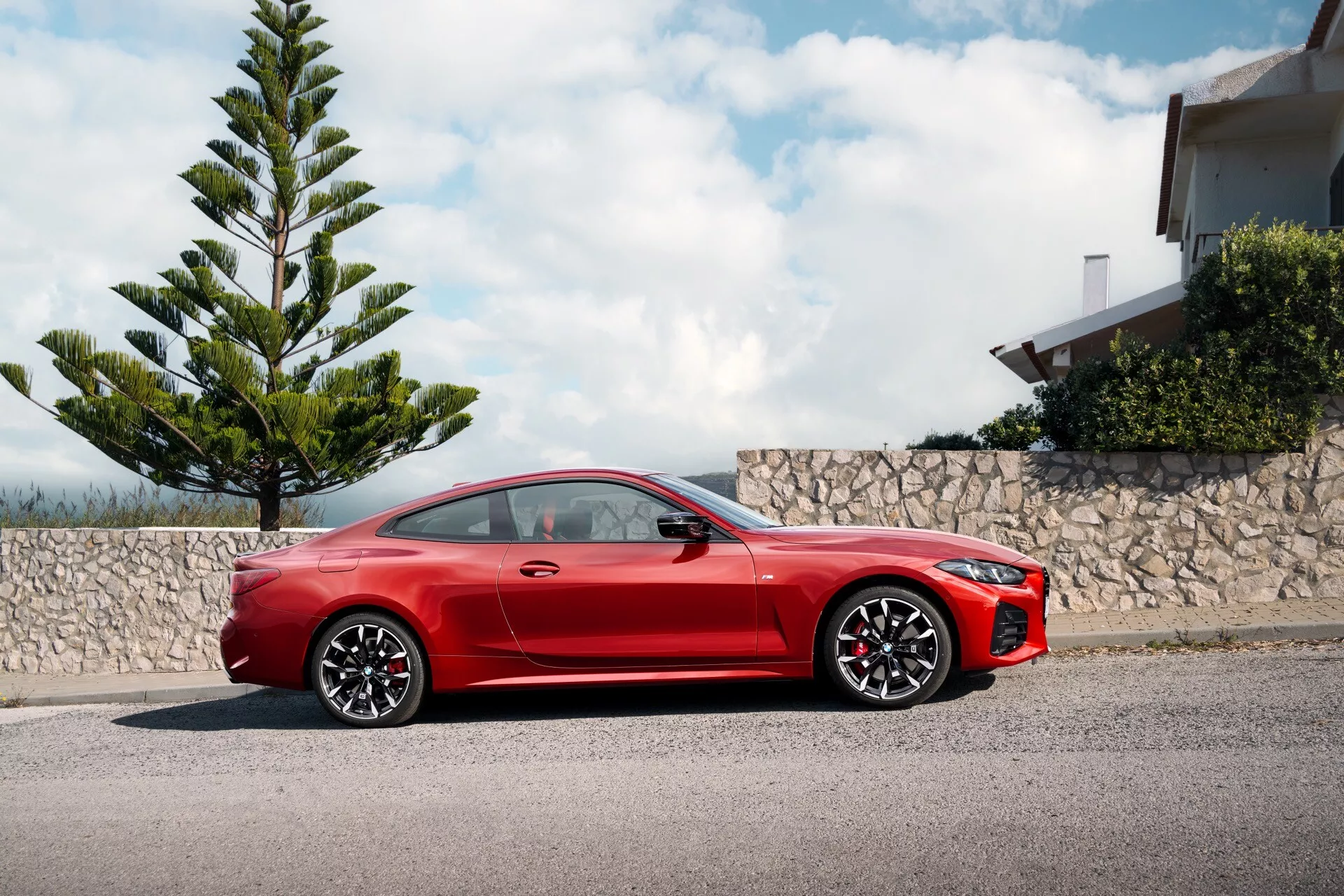 Rdeči športni kupe BMW serije 4 2024 (G22-G23-G26-G82-G83-Facelift) parkiran ob robniku, prikazuje celoten stranski profil z dinamično perspektivo iz nizkega kota, ki poudarja njegov športni dizajn, dolg pokrov motorja, elegantno linijo strehe, velika lita kolesa, aerodinamična stranska ogledala, ročaje vrat poravnane s karoserijo, zmogljiv zavorni sistem z rdečimi zavornimi čeljustmi, LED žaromete in agresiven sprednji odbijač, pod delno oblačnim nebom ob zelenju in stanovanjski zgradbi