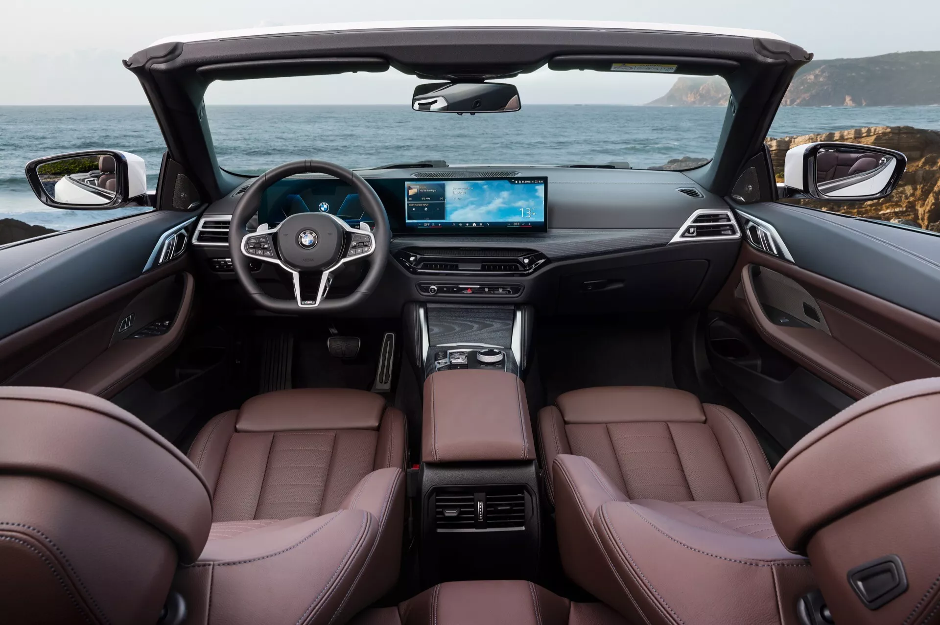 Notranjost kabrioleta BMW serije 4 za leto 2024 prikazuje rjavo usnjene sprednje sedeže, volan s tremi kraki in integriranimi kontrolniki, digitalni širokozaslonski zaslon armaturne plošče in elegantno sredinsko konzolo v razkošni kabini s spuščeno zložljivo streho, prikazuje slikovito obalno ozadje