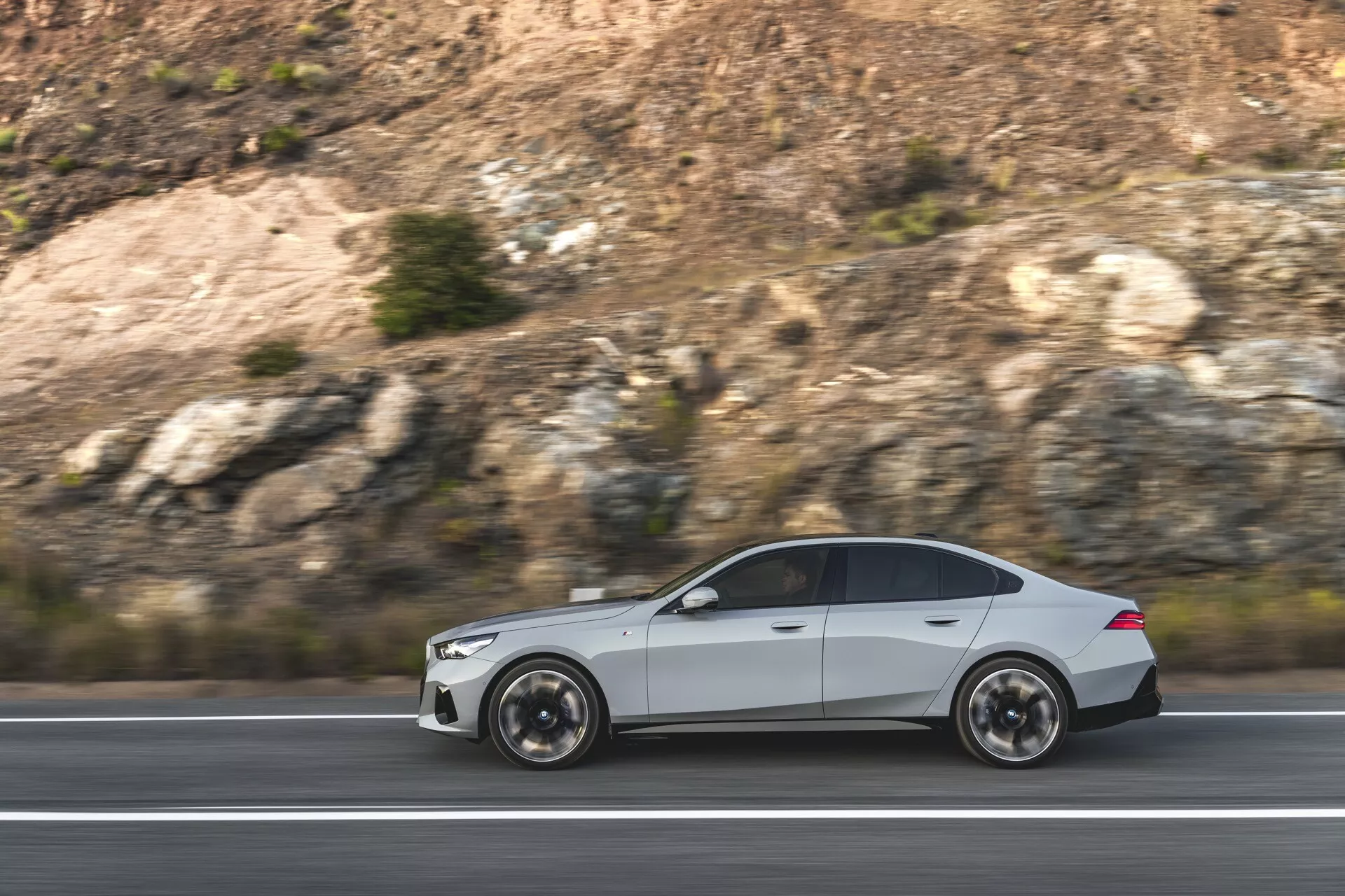 BMW serije 5 (G60-G61) letnik 2023 v gibanju, ki prikazuje svojo aerodinamično obliko in športno estetiko z vidnimi večkrakimi kolesi in elegantnimi obrisi karoserije, posneto s strani na avtocesti