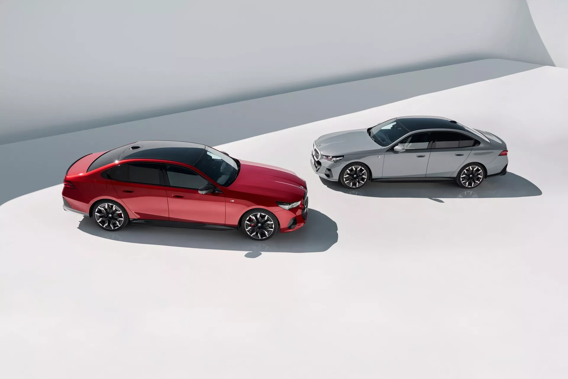 Pogled z višine na BMW serije 5 (G60-G61) letnik 2023, ki prikazuje model v živo rdeči in srebrni barvi na enostavni površini, poudarja zgornje in stranske profile, platišča iz lahkih zlitin z večimi kraki in elegantne oblikovalske lastnosti iz visokega kota