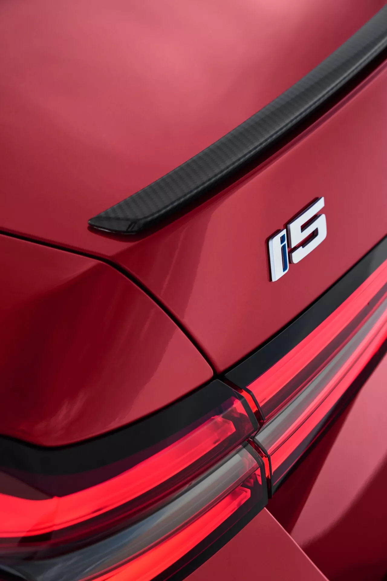 Podrobnost desnega zadnjega kota BMW serije 5 (G60-G61) letnik 2023 z integriranim spojlerjem na pokrovu prtljažnika, oznako i5, sklopom LED zadnjih luči in sijočo metalik rdečo barvo