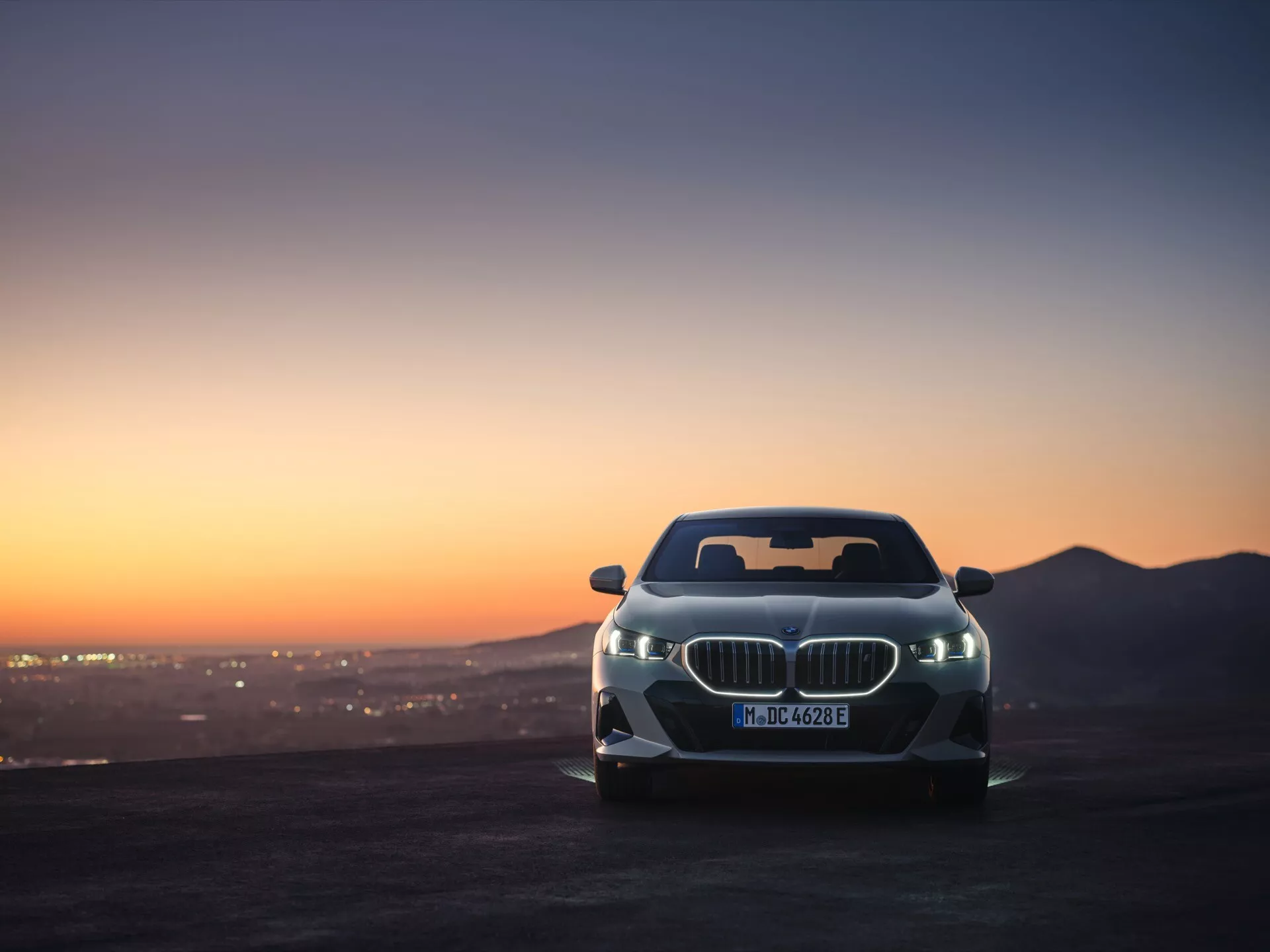 Poševni sprednji pogled na BMW serije 5 (G60-G61) 2023 s kultno ledvično masko in naprednimi LED žarometi med slikovitim sončnim vzhodom ali zahodom, ki prikazuje aerodinamično obliko vozila in luksuzno estetiko ob ozadju gorske pokrajine in svetlečega mestnega krajine.