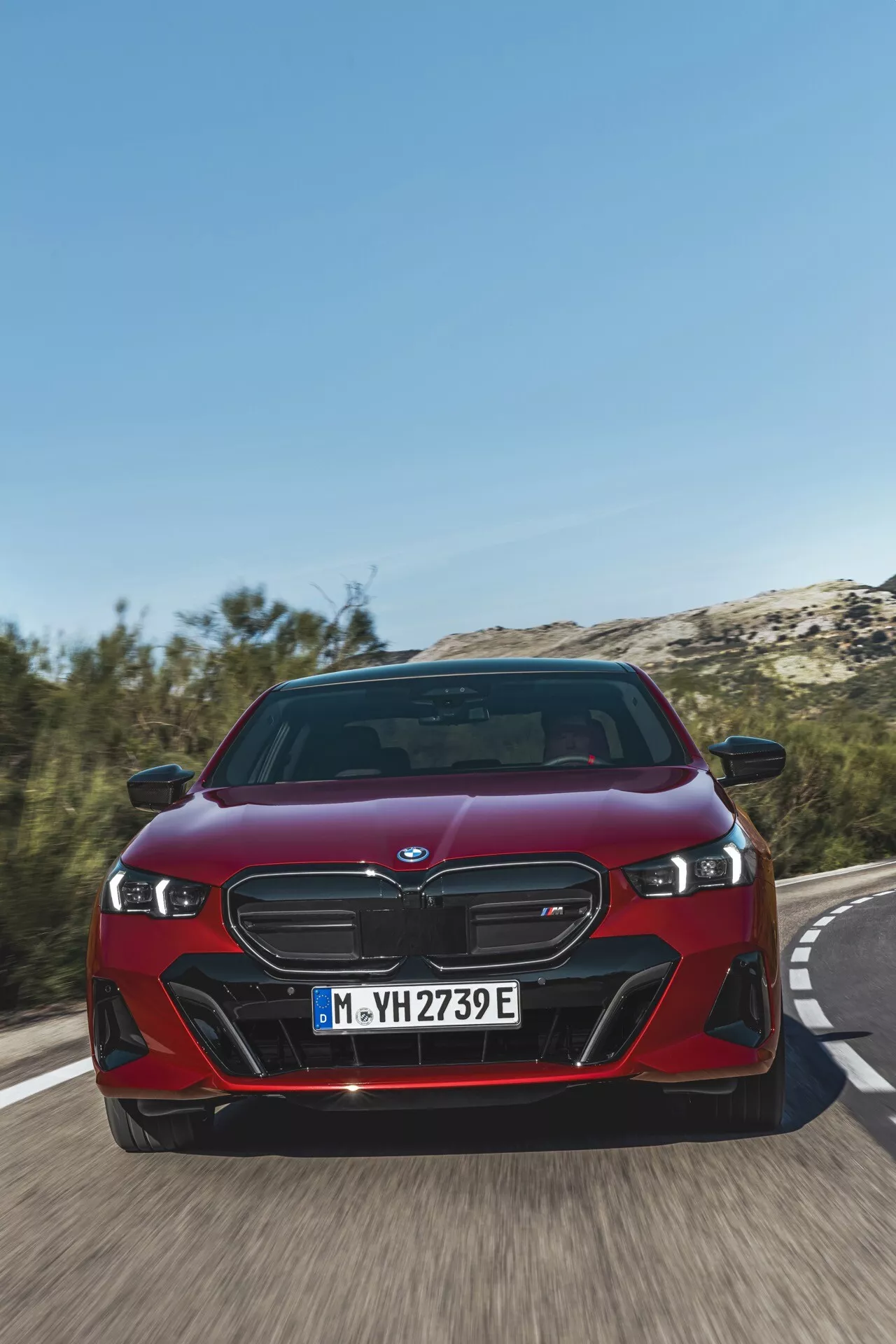 BMW serija 5 (G60-G61) letnik 2023 v rdeči barvi, prikazuje svojo značilno ledvičasto rešetko, LED žaromete in dinamični sprednji odbijač iz nizkega sprednjega kota na cesti, poudarja hitrost vozila in napredne elektrificirane funkcije iPerformance