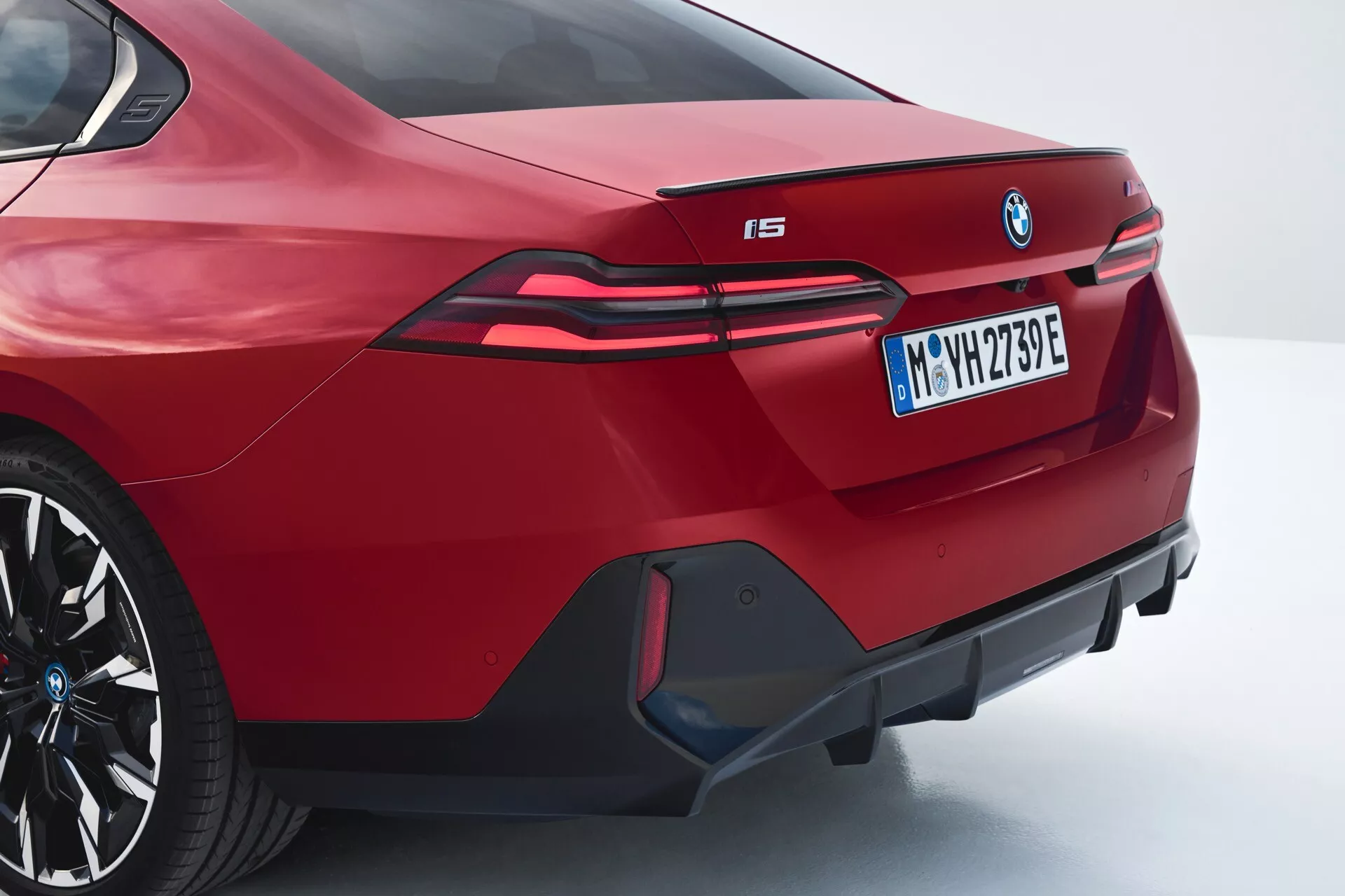 Zadnji kotni pogled na rdeč BMW i5 letnik 2023, ki prikazuje športno zasnovo sedana, integriran spojler, izstopajoči znak BMW, podaljšane LED zadnje luči, ki segajo v zadnja krila, oznako električnega modela 'i5', agresivni zadnji odbijač z dekorativnim difuzorjem in lažnimi izpušnimi luknjami, v tričetrtinskem zadnjem in stranskem profilu.