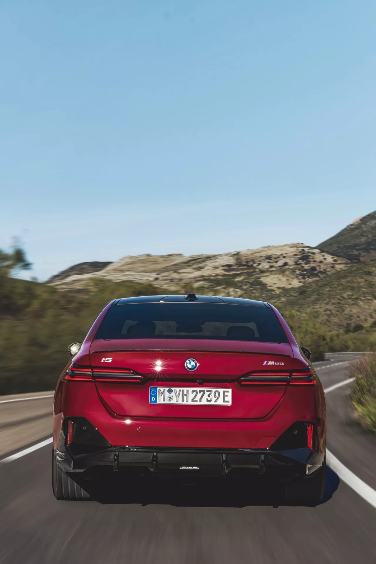 Zadnji pogled na živo rdeči sedan BMW serije 5 (G60-G61), ki se vozi po cesti, z LED zadnjimi lučmi v obliki črke L, dvojnim izpušnim sistemom, znakom eDrive in senzorji za parkiranje pod jasno dnevno svetlobo.