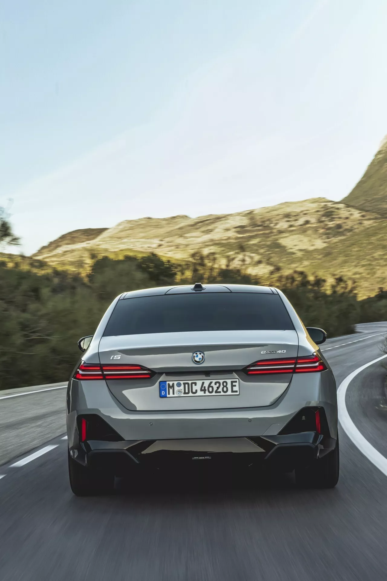 Zadnji pogled na BMW serije 5 (G60-G61) 2023, ki prikazuje LED zadnje luči, senzorje za pomoč pri parkiranju in oznako modela električno-hibridnega vozila v dinamičnem cestnem okolju