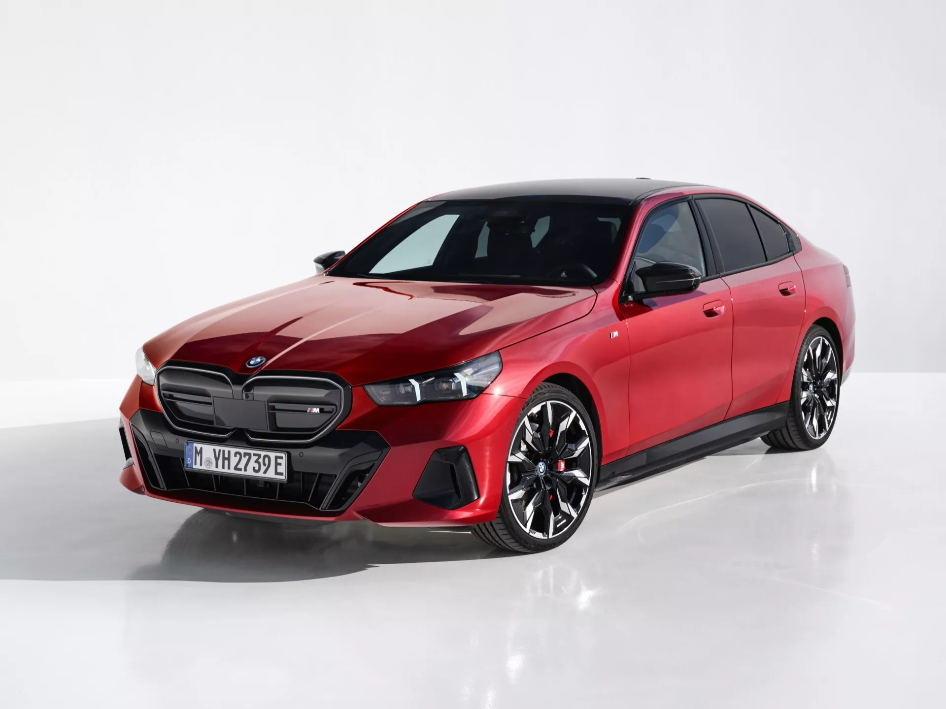 Rdeča štirivratna limuzina BMW serije 5 (G60-G61) letnik 2023 iz perspektive tri četrt spredaj, ki prikazuje oblikovanje rešetke, vitke žaromete in kiparski stranski profil na odsevni površini z belim ozadjem