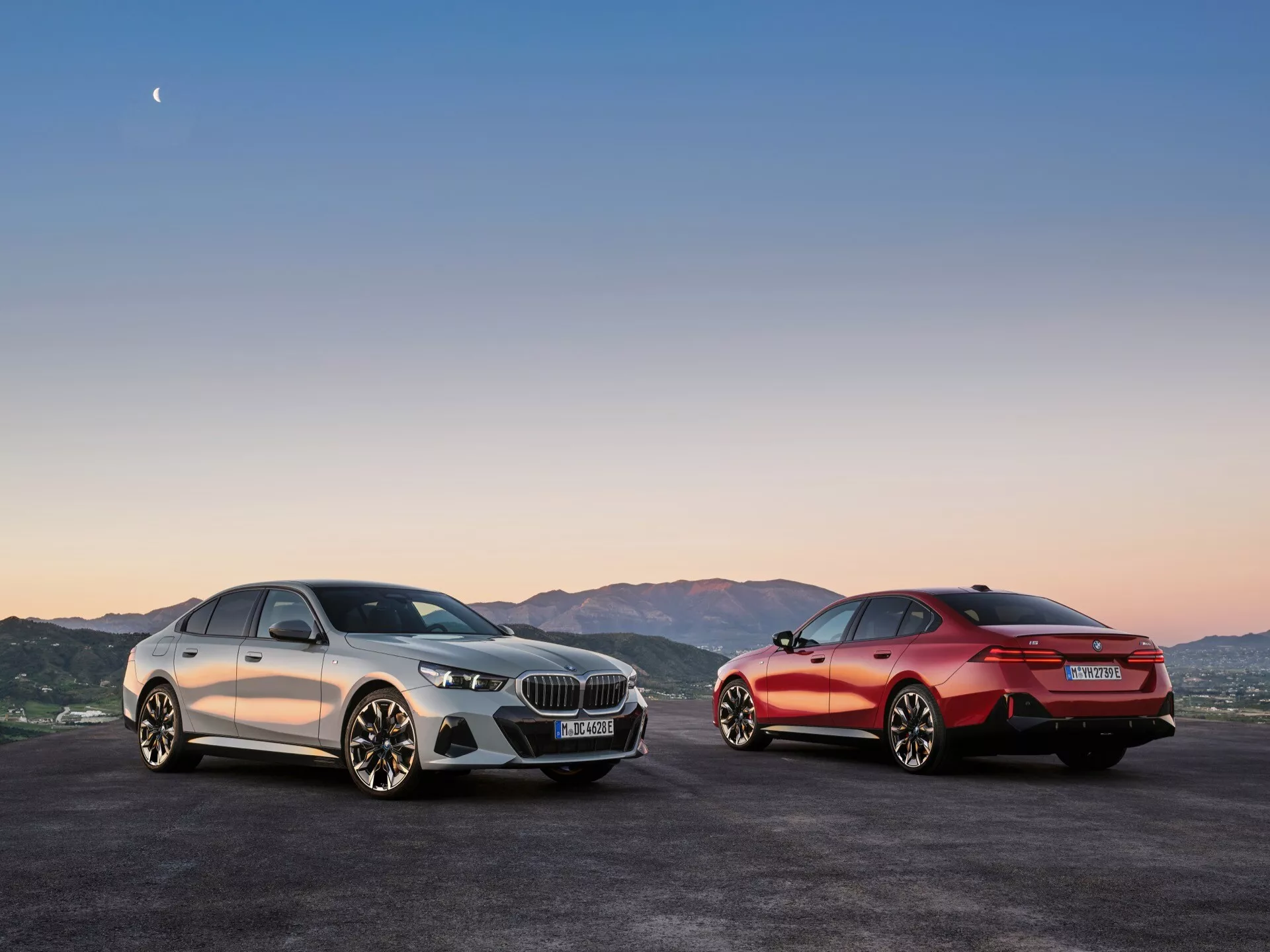 BMW serije 5 (G60-G61) 2023 predstavljen v zlati uri z ambientalno razsvetljavo ob sončnem zahodu, z dvema modeloma v kontrastnih pogledih proti panoramskim hribom, poudarjajoč športno a hkrati razkošno oblikovanje in napredne tehnološke funkcije vozil.