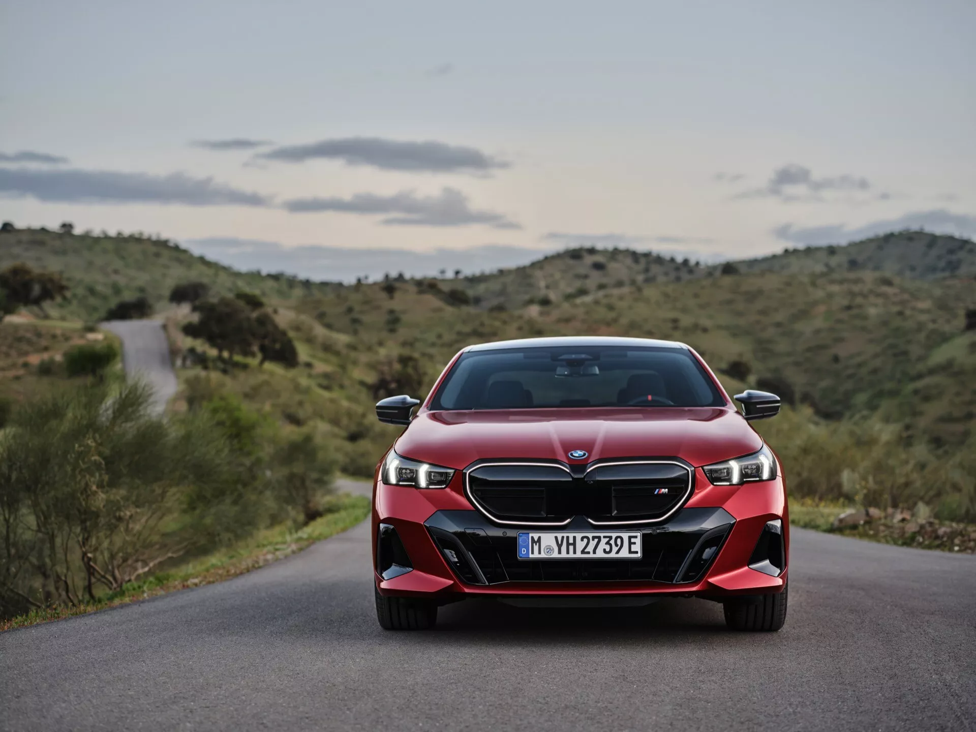 Sprednji pogled na rdeči BMW serije 5 (G60-G61) letnik 2023, z LED žarometi, dvojno ledvično masko in športnim odbijačem proti ozadju ovinkaste ceste in gričevnate pokrajine