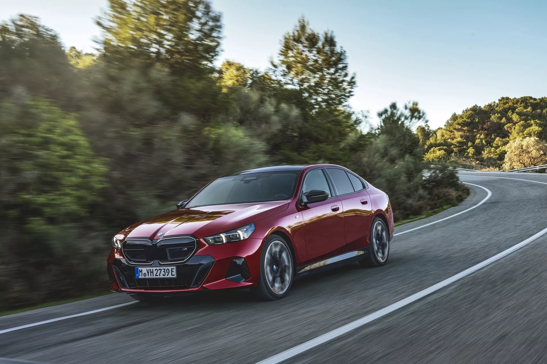Dinamičen tričetrtinski pogled na BMW serije 5 (G60-G61) iz leta 2023 v gibanju na ukrivljeni cesti sredi podeželske pokrajine, poudarja njegovo metalik rdečo barvo, značilno sprednjo rešetko, elegantne LED žaromete, aerodinamična stranska ogledala, športne linije, pnevmatike z nizkim profilom in večkraka lita kolesa