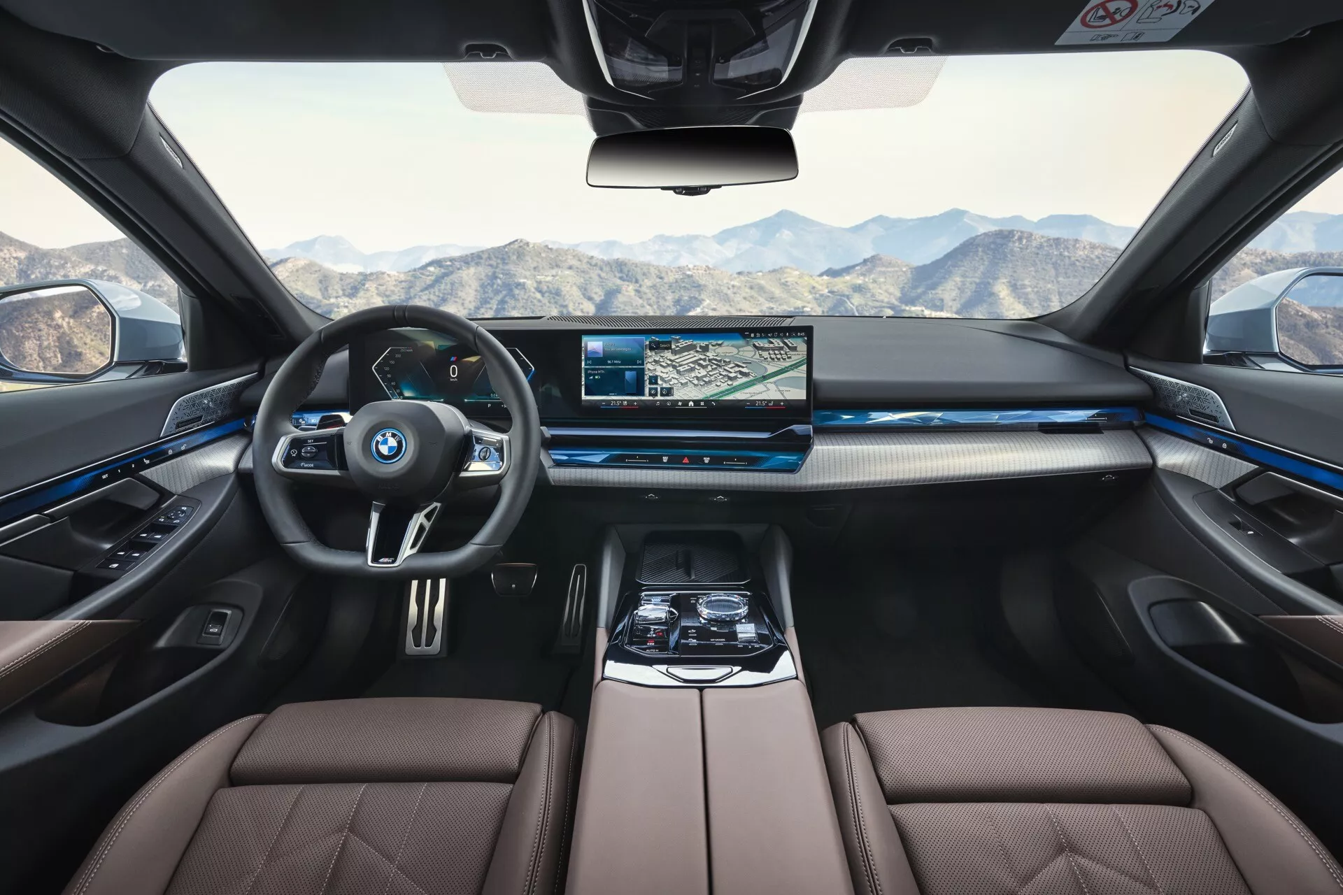 Notranjost BMW serije 5 (G60-G61) letnik 2023 iz perspektive zadnjega sedeža, ki prikazuje armaturno ploščo, volan, sredinsko konzolo in predel za potnike spredaj z luksuznimi sedeži v dveh tonih in modro ambientalno osvetljavo