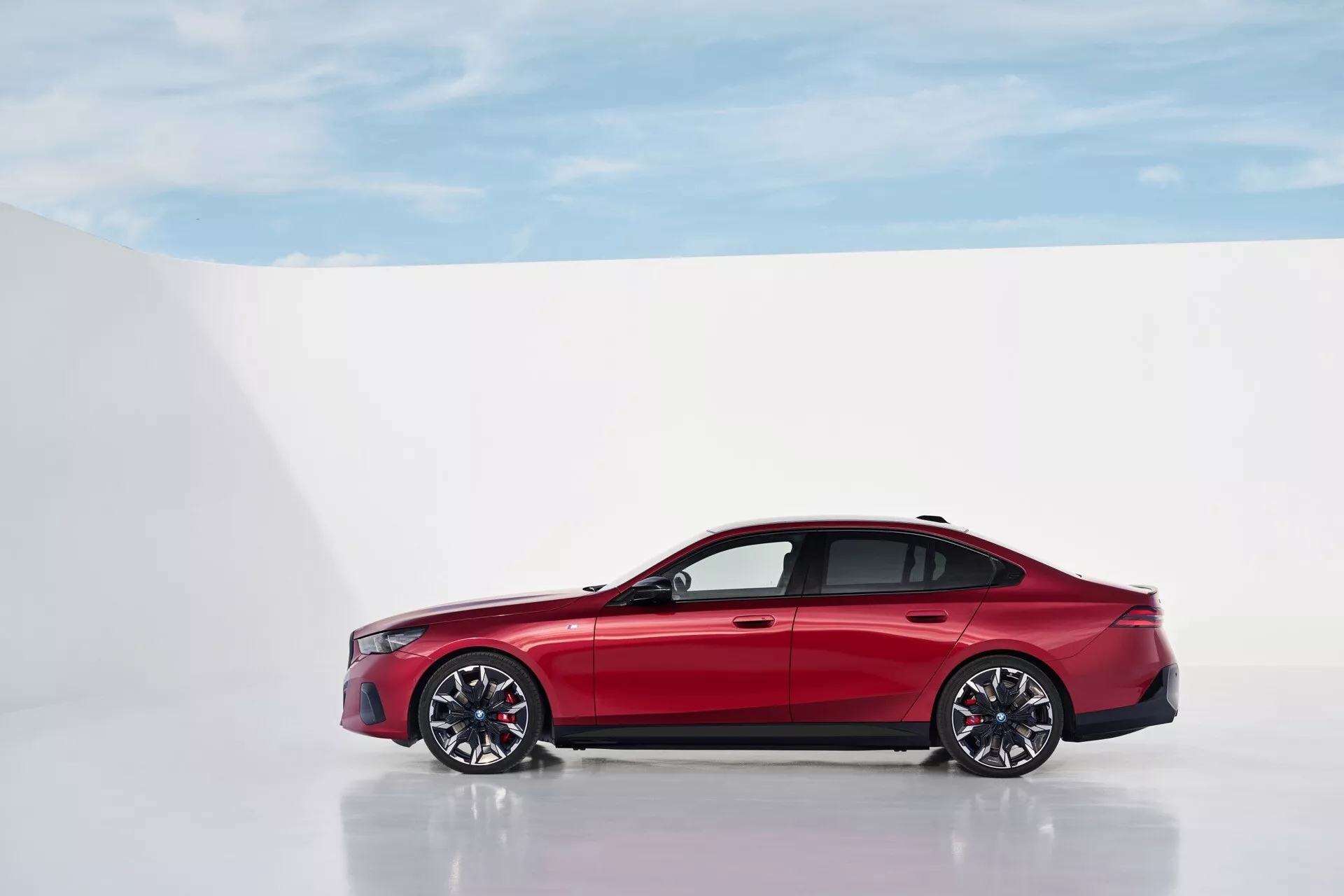 BMW serija 5 (G60-G61) letnik 2023 v živahni rdeči barvi, posnet iz stranskega profila na odsevni površini proti beli ozadju, poudarja njegov eleganten dizajn in aerodinamične lastnosti, vključno z večkrakimi aluminijastimi kolesi in pnevmatikami z nizkim profilom.