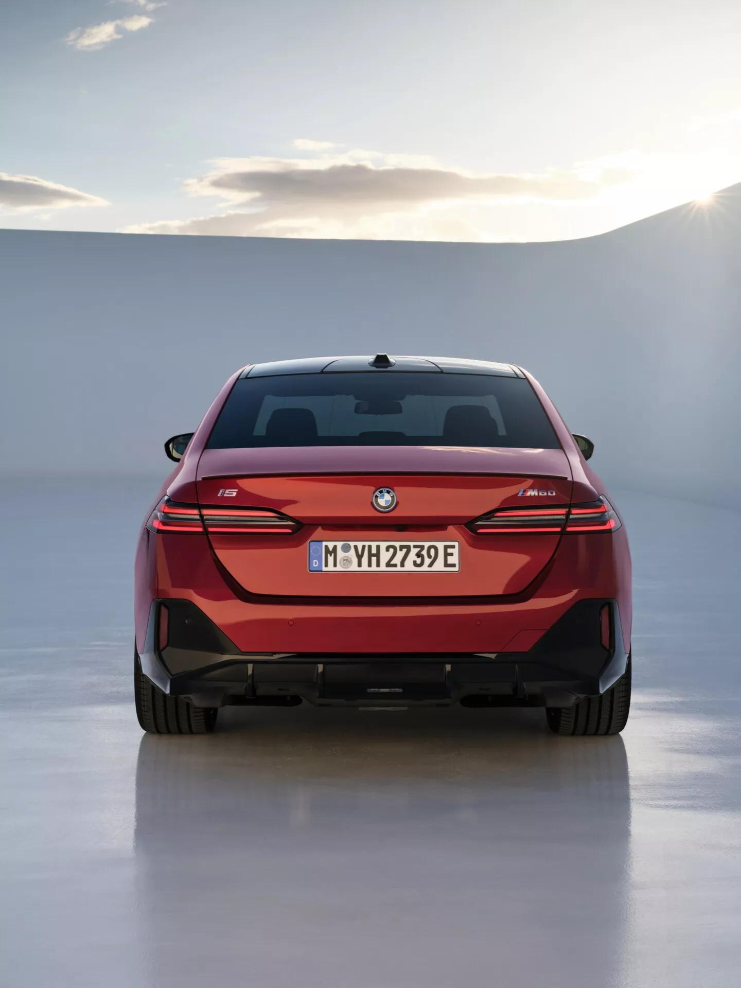 Zadnji pogled na BMW serije 5 (G60-G61) 2023 z rdečo zunanjostjo, elegantnimi horizontalnimi LED zadnjimi lučmi in oznako električnega modela, proti ozadju z mehko obzorje in studijsko razsvetljavo ali oblačnim nebom