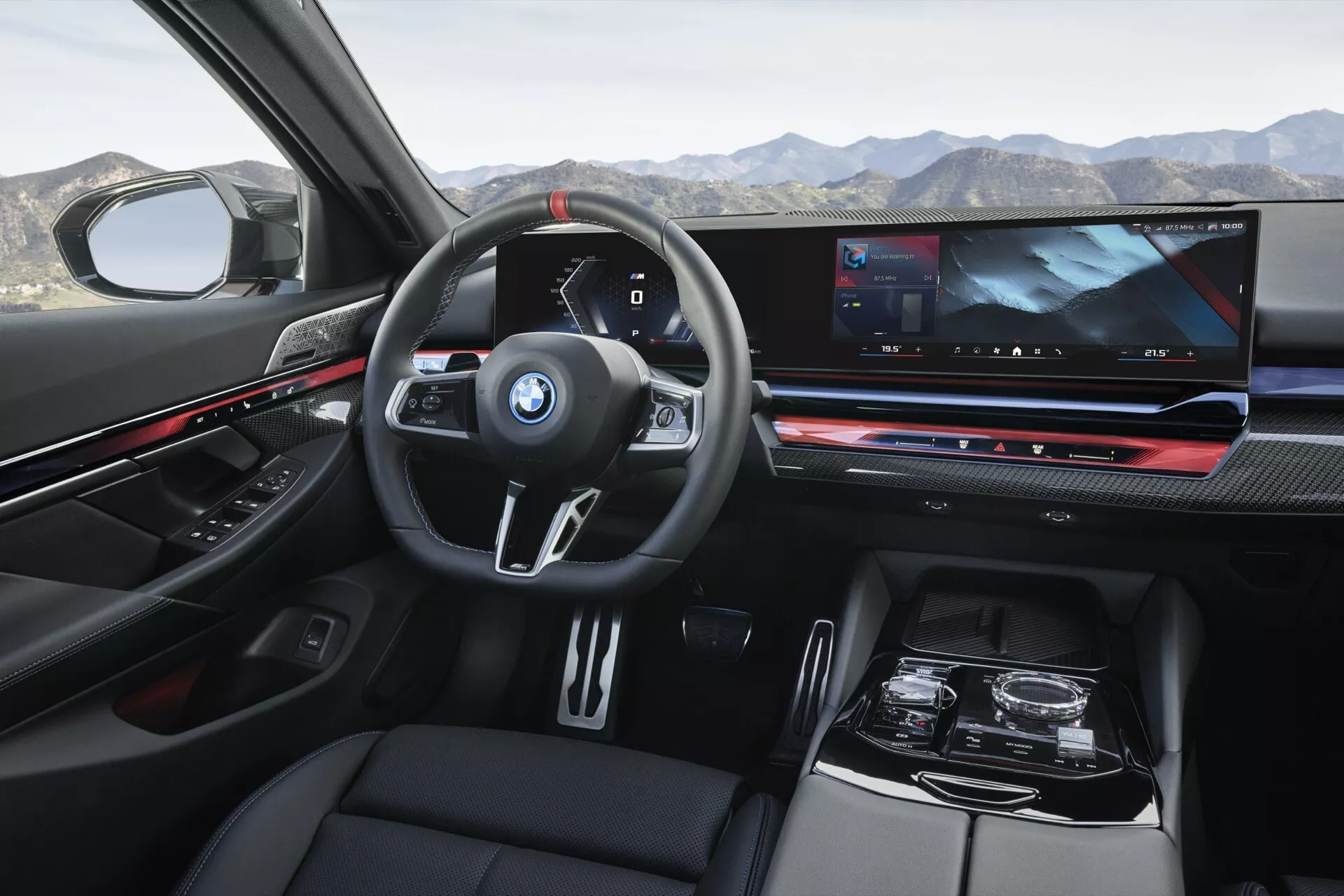 Pogled na notranjost BMW serije 5 (G60-G61) letnik 2023 iz perspektive zadnjega potnika prikazuje armaturo, večnamenski volan z logotipom BMW, digitalni instrumentni zaslon, obsežen zaslon za informacije in zabavo, elektronsko prestavno ročico in razkošne materiale vključno z usnjem in detajli iz ogljikovih vlaken.