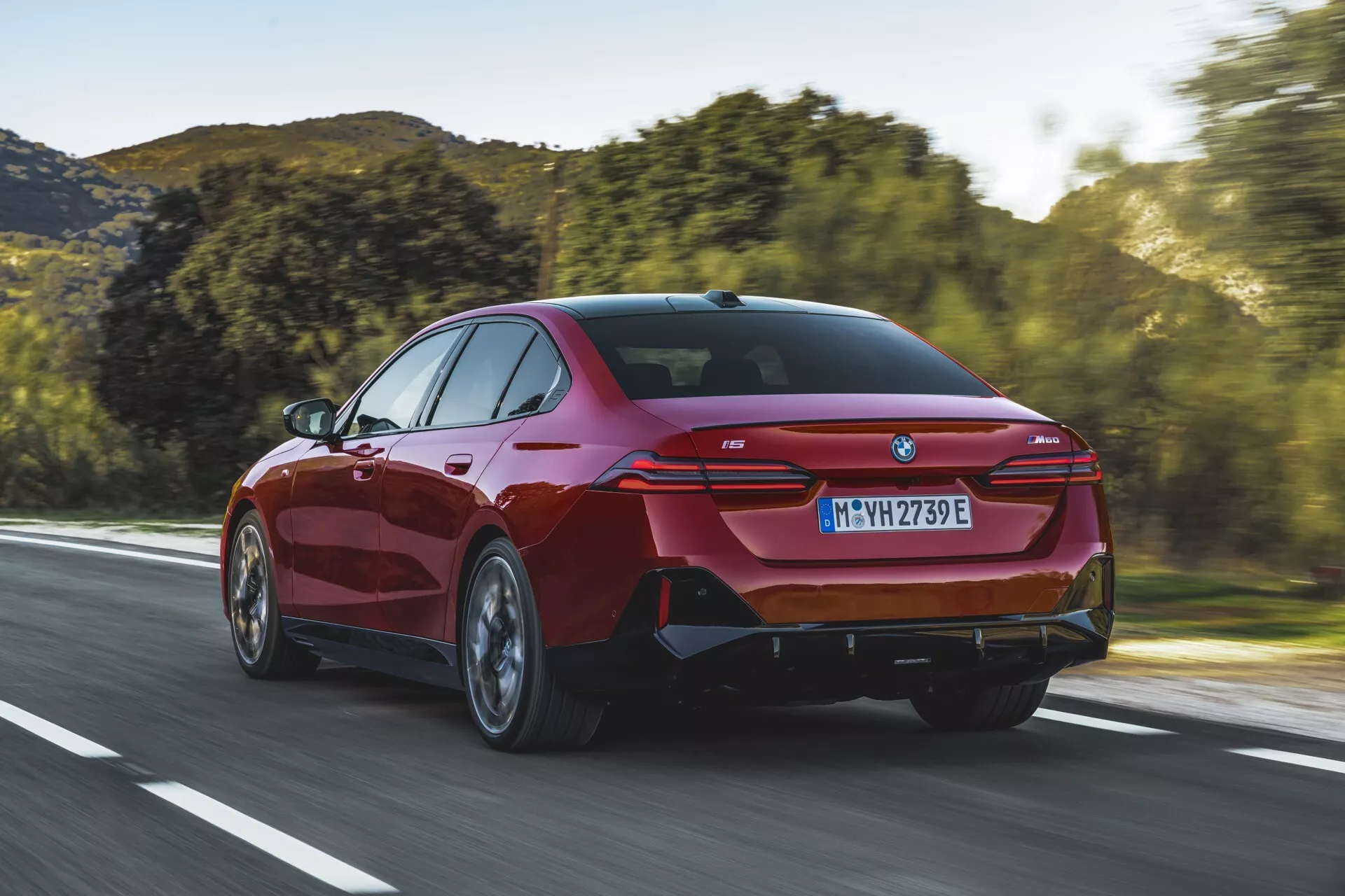 Moderen sedan BMW serije 5 (G60-G61) iz leta 2023, vozeč se po odprti cesti, prikazan iz treh četrt zadnjega pogleda, ki razkriva elegantne zadnje luči, športni zadnji odbijač z integriranimi izpušnimi priključki in značilno coupé-podobno silhueto.