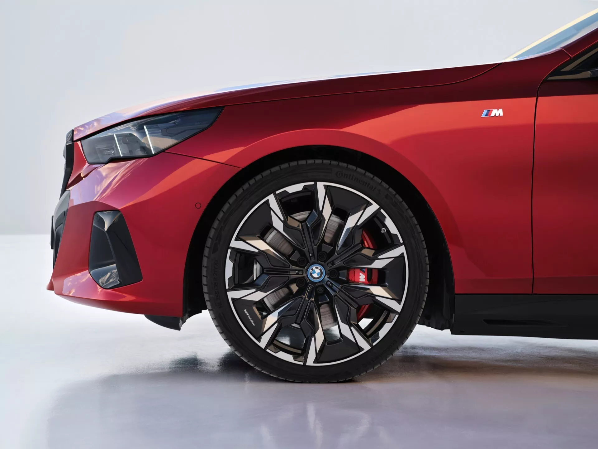 Podroben pogled na sprednji levi del BMW serije 5 (G60-G61) letnik 2023, prikazujemoč kolo in blatnik, z detajli, kot so logotip na modrem središču kolesa, rdeč zavorni čeljust in na zmogljivost usmerjen sprednji odbijač.