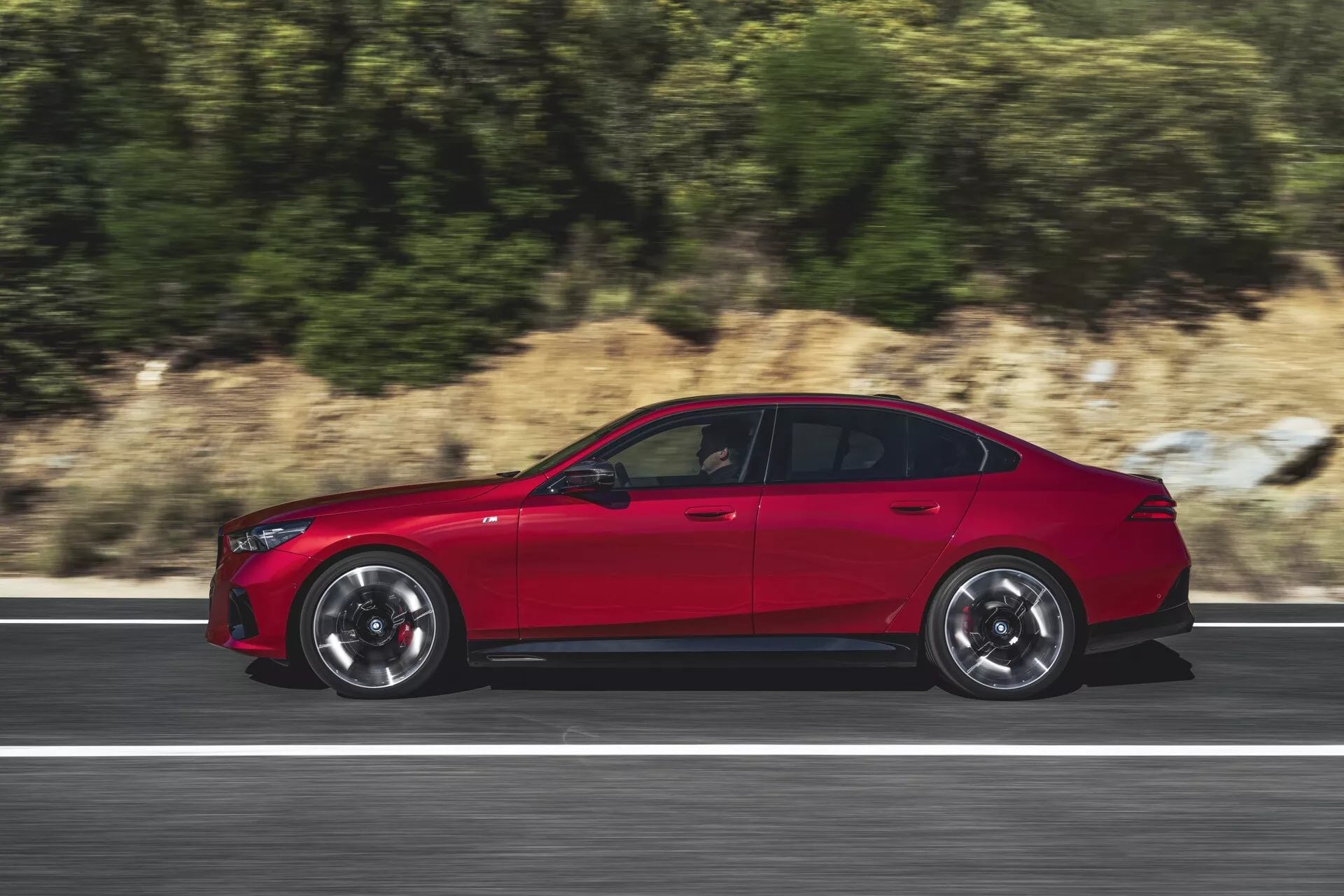 Dinamični stranski profil BMW serije 5 (G60-G61) leta 2023 v gibanju na cesti, ki poudarja športno oblikovanje, velika platišča iz zlitine z več kraki in svetlečo rdečo barvo, ki poudarja aerodinamiko in napredne tehnološke funkcije