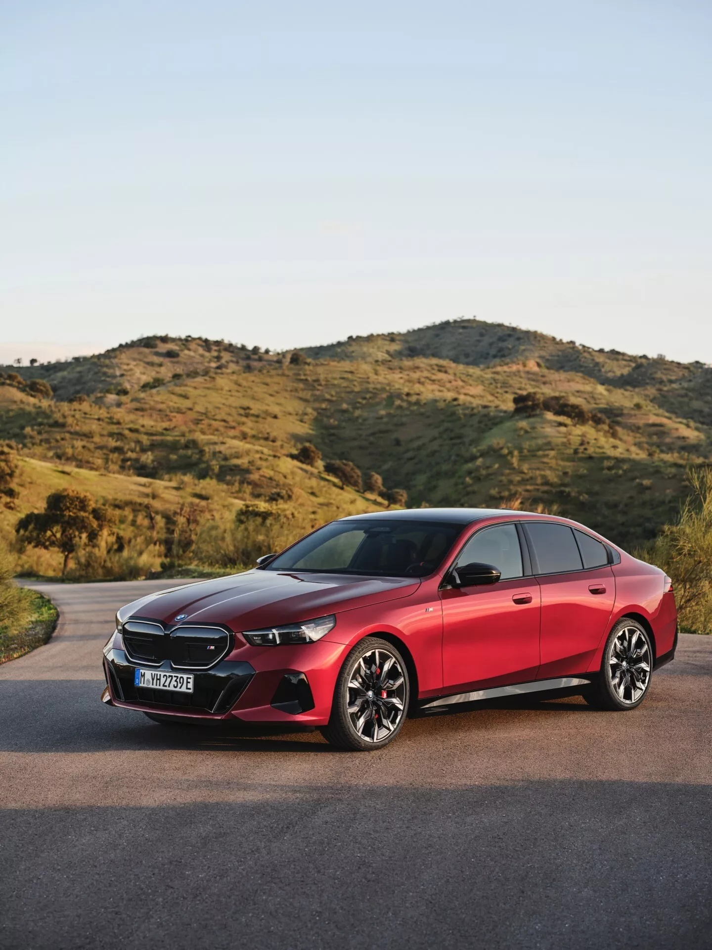 BMW serije 5 2023 (G60-G61) tričetrtinski sprednji pogled na asfaltni cesti z naravnim, hribovitim ozadjem, prikazuje LED žaromete, senzorje, integrirane v rešetko, zračne reže, športna platišča iz lahkih zlitin, sistem za ključavničenje brez ključa in visokokakovostni sijajni zaključek
