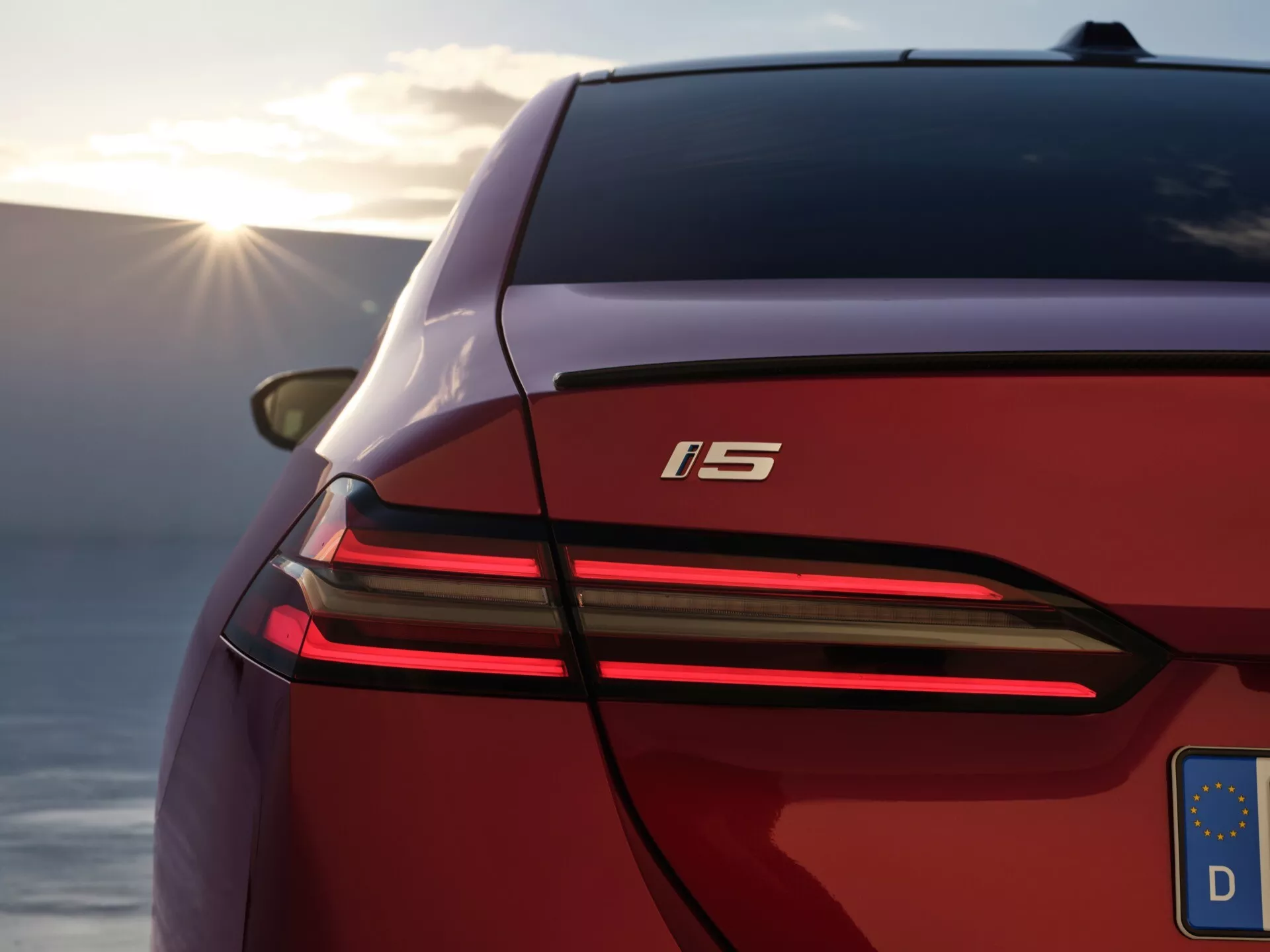 Posnetek od blizu, ki prikazuje zadnji levi del BMW serije 5 (G60-G61) letnik 2023, poudarja njegovo elegantno ukrivljenost karoserije, značilno celotno širino zavornih luči, sodobno zasnovo LED zadnjih luči in oznako 'i5' proti zamegljenemu naravnemu ozadju s toplim sončnim sijem.