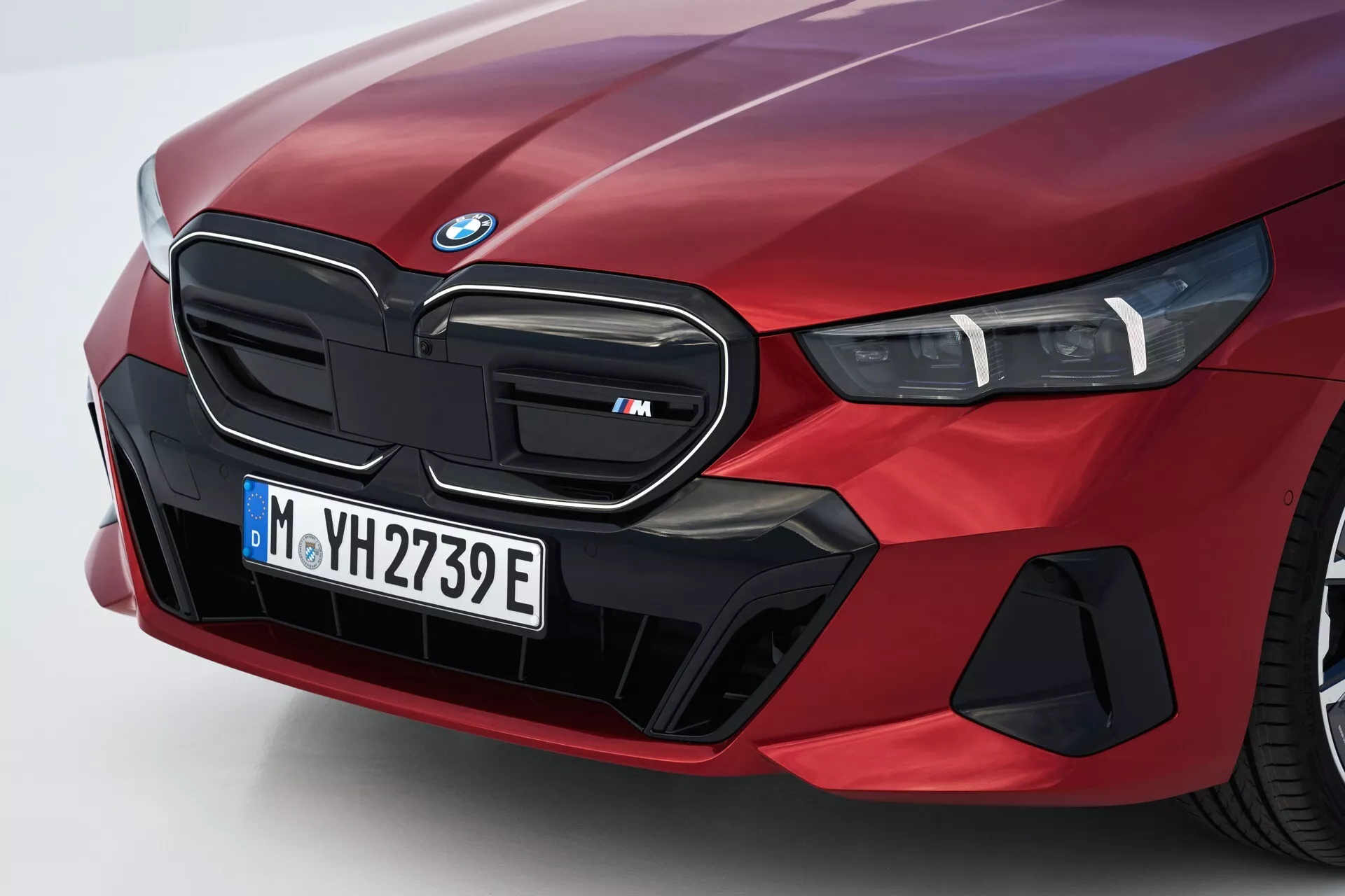 Podroben kotni pogled na sprednji levi del rdečega BMW serije 5 (G60-G61) letnik 2023, z ikonično ledvično masko označeno kot model 'M', LED žarometi in specializirani aerodinamični elementi, ki nakazujejo električne ali hibridne zmožnosti