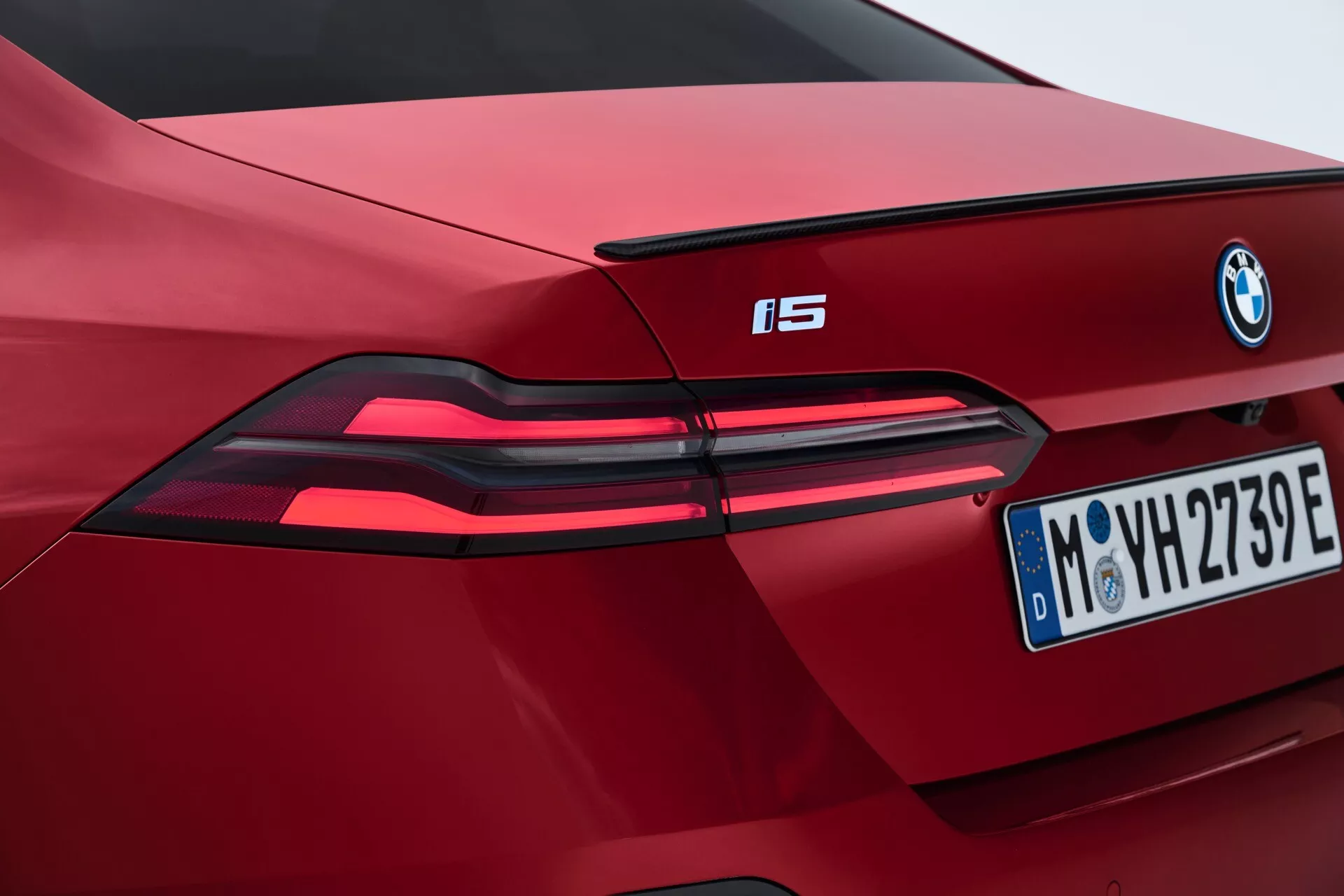 Podroben pogled na delni zadnji del rdečega BMW i5 letnik 2023, ki prikazuje sodobne LED zadnje luči, izstopajoči znak BMW in evropsko registrsko tablico iz Münchna, poudarjajoč značilnosti električnega vozila