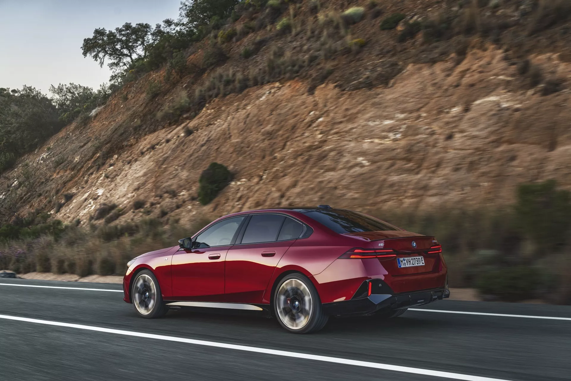 Rdeča štirivratna limuzina BMW serije 5 (G60-G61) v dinamičnem gibanju na cesti, posneta iz stranskega zadnjega zornega kota, ki poudarja aerodinamično oblikovanje avtomobila, LED zadnje luči, zadnje kolo in odbijač