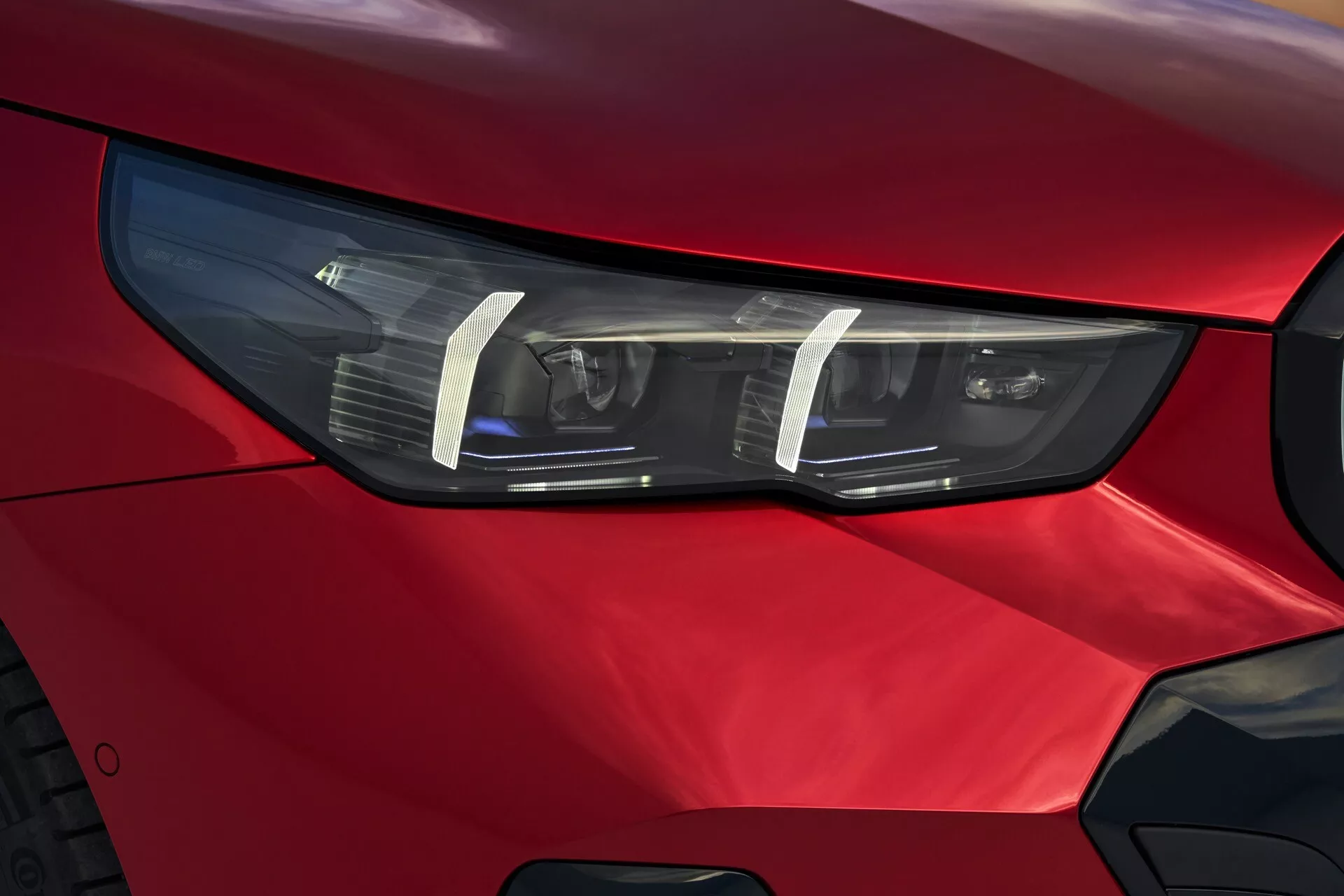 Pogled od blizu na žaromet na voznikovi strani BMW serije 5 (G60-G61) 2023 z LED ali HID tehnologijo, vitkim kotnim dizajnom in živahno rdečo barvo, ki poudarja napredno avtomobilsko oblikovanje in premium estetiko.