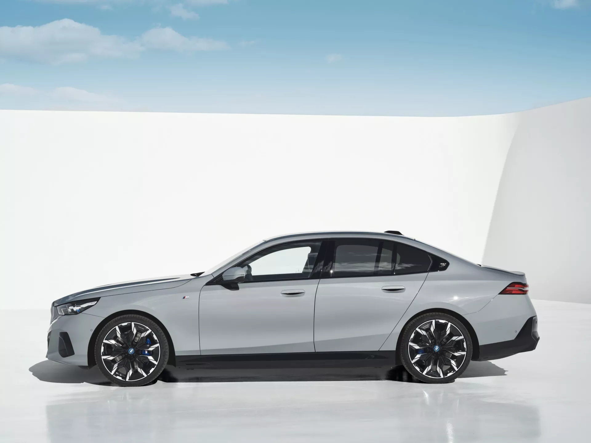 Bočni pogled na BMW serije 5 (G60-G61) 2023 v simuliranem minimalističnem okolju, ki prikazuje njegovo športno limuzinsko silhueto v svetlo sivi barvi z velikimi zlitimi platišči in elegantnim dizajnom
