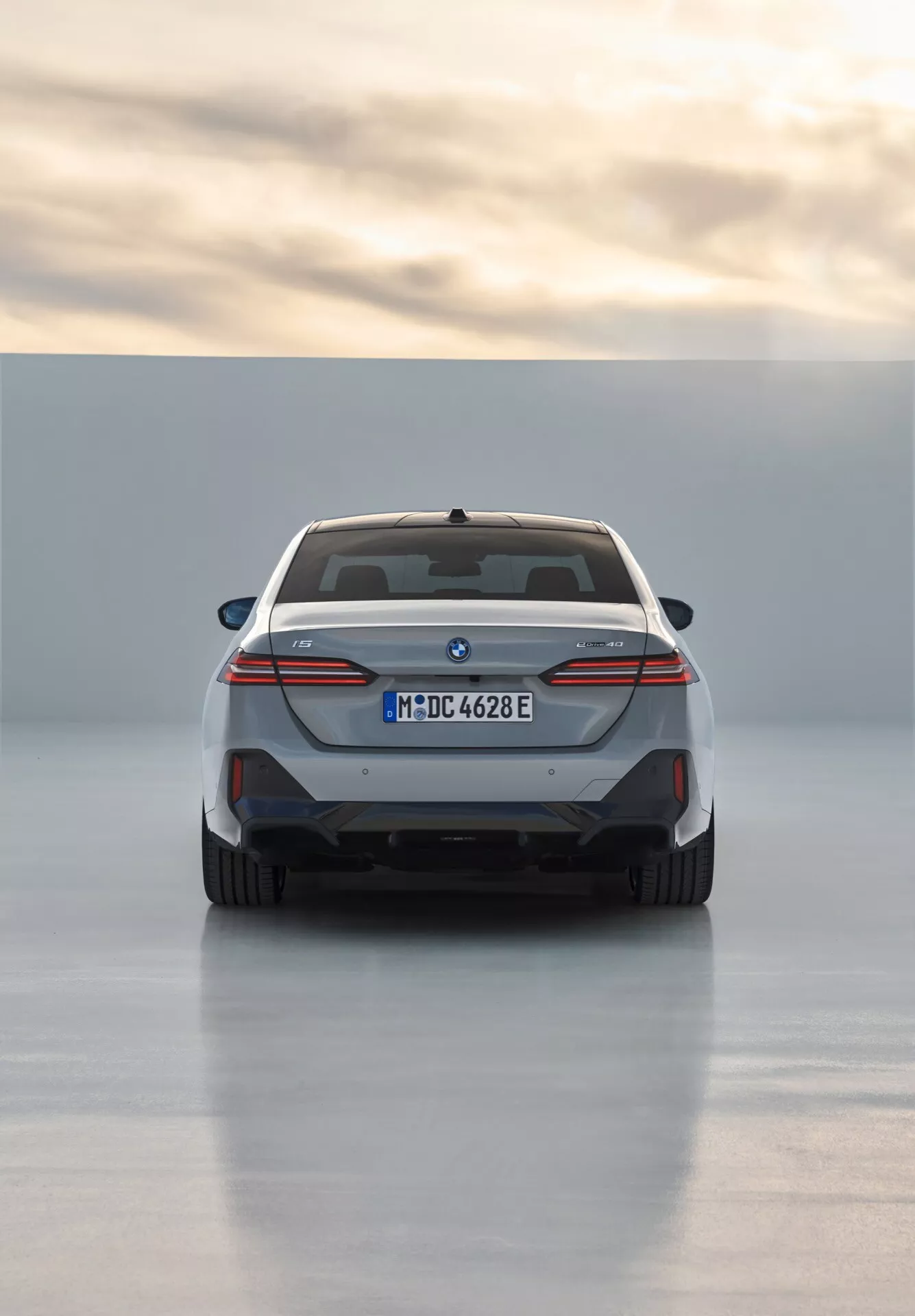 Zadnji pogled na BMW serije 5 (G60-G61) 2023 s polnoširinskim dizajnom zadnjih luči, modrimi poudarki električnega modela, dvojnimi izpuhi in športnim zadnjim difuzorjem, registriranim v Münchnu z učinkom odsevnega talnega osvetlitve