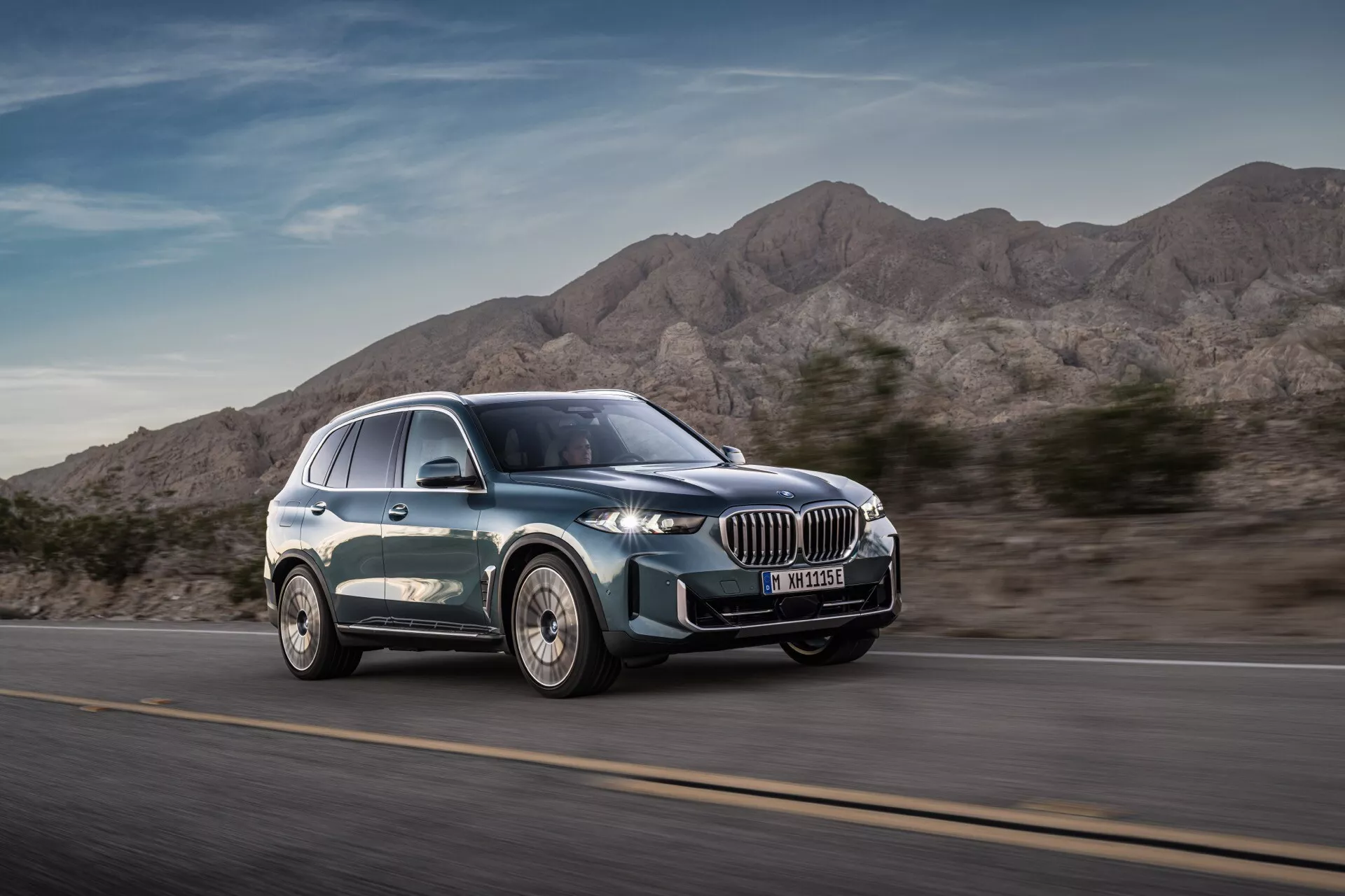 Sodobni luksuzni SUV BMW X5 2023 (G05-F95 Facelift) v gibanju iz sprednjega pogleda tri četrtine poudarja dinamično oblikovanje, veliko ledvičasto masko, ozke LED žaromete, lita kolesa z večkrakim dizajnom in gladko silhueto z metalik barvo