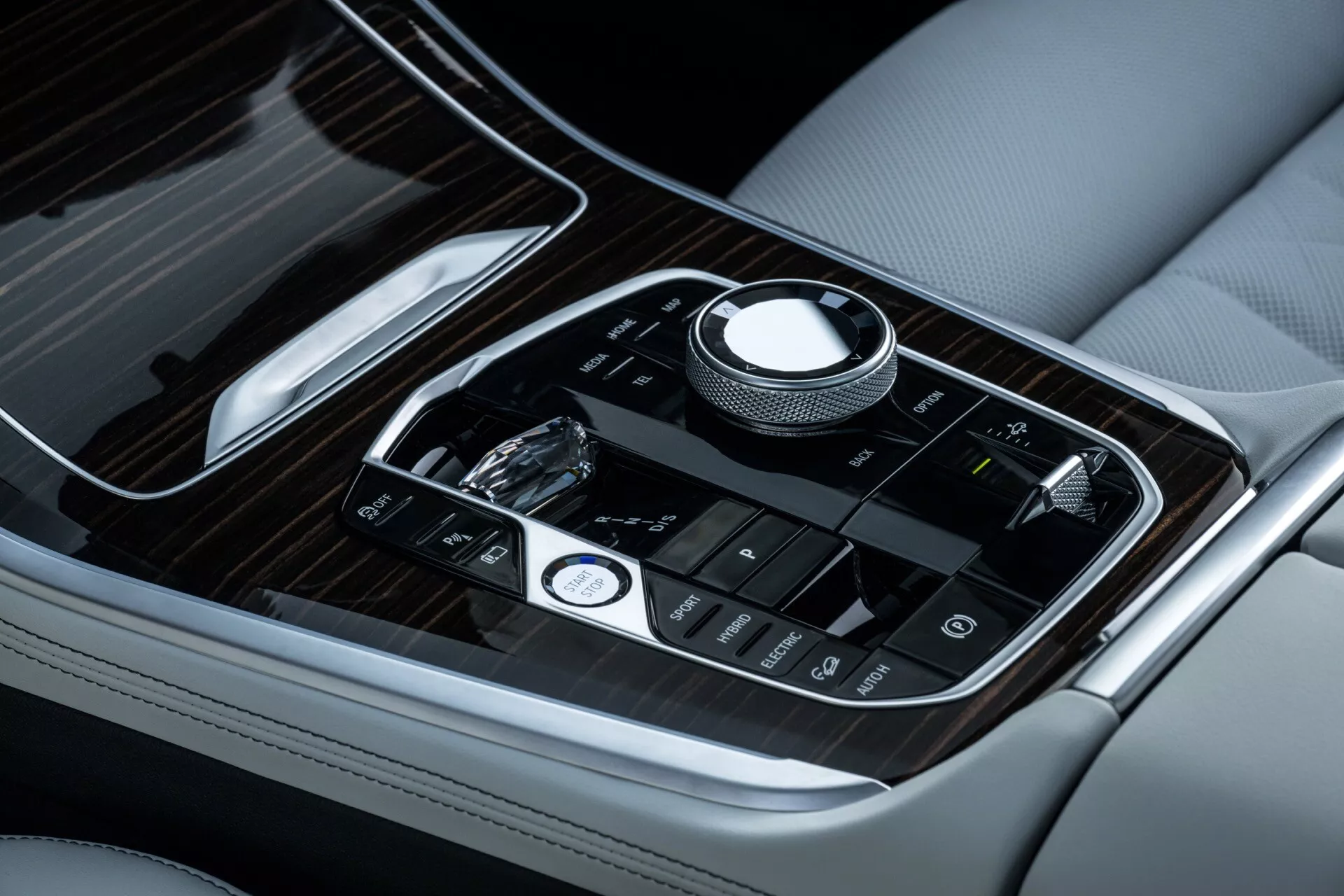 Približek sredinske konzole BMW X5 2023 (G05-F95 Facelift), ki prikazuje luksuzno usnje s kontrastnim šivanjem, leseni vložki in napredne občutljive na dotik kontrole z rotacijskim gumbom za navigacijo po multimedijskem sistemu