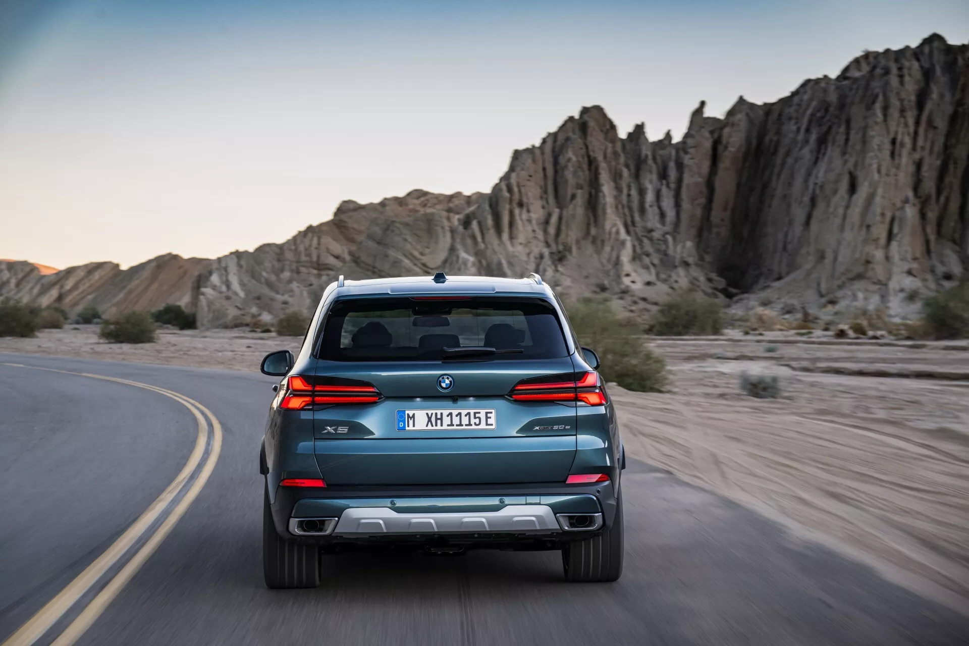 Zadnji pogled na BMW X5 G05 F95 facelift 2023, vožnjo po ovinkasti poti, s elegantnimi linijami, dvojnim izpuhom in parkirnimi senzorji na ozadju pustolovske skalnate pokrajine