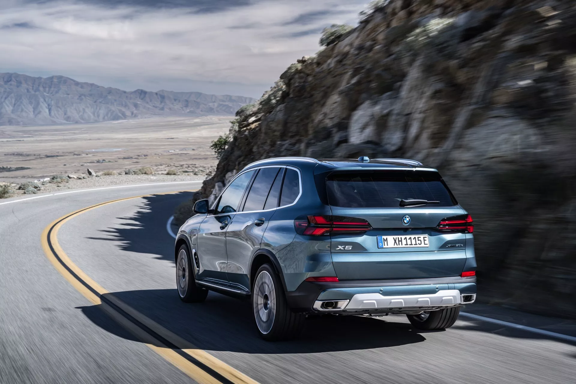 Dinamični zadnji tričetrtinski pogled na BMW X5 2023 (G05-F95 Facelift) na ovinkasti gorski cesti, ki prikazuje elegantno zadnje steklo, športni spojler in luksuzna lita kolesa