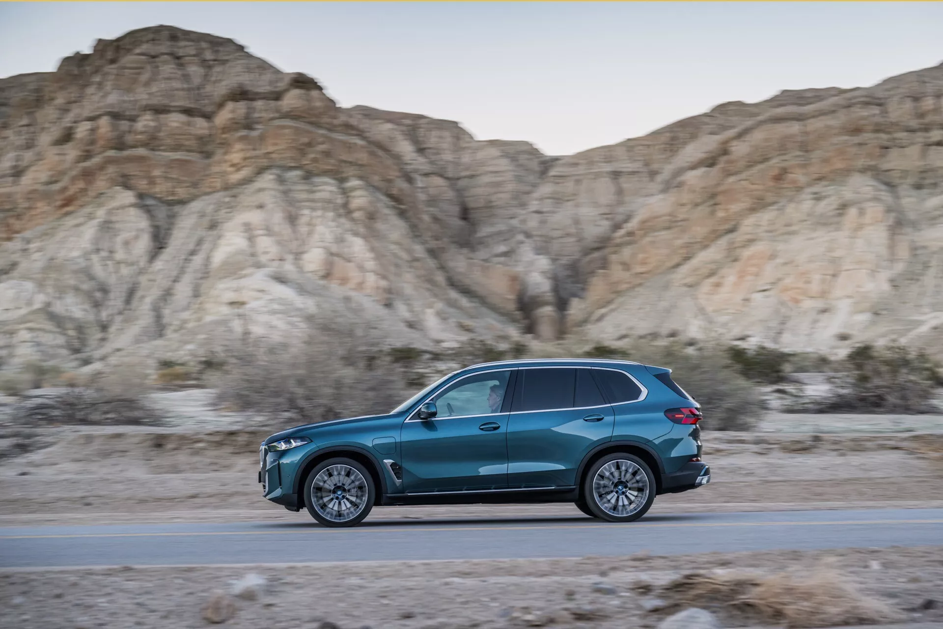 Dinamični stranski profil BMW X5 2023 (G05-F95 prenova) v gibanju, modri SUV v obliki solze na cesti z ozadjem skalnatih, pustih hribov med zlato uro, prikazuje elegantno oblikovanje, aerodinamično učinkovitost, večkraka zlitinska kolesa in LED osvetlitev