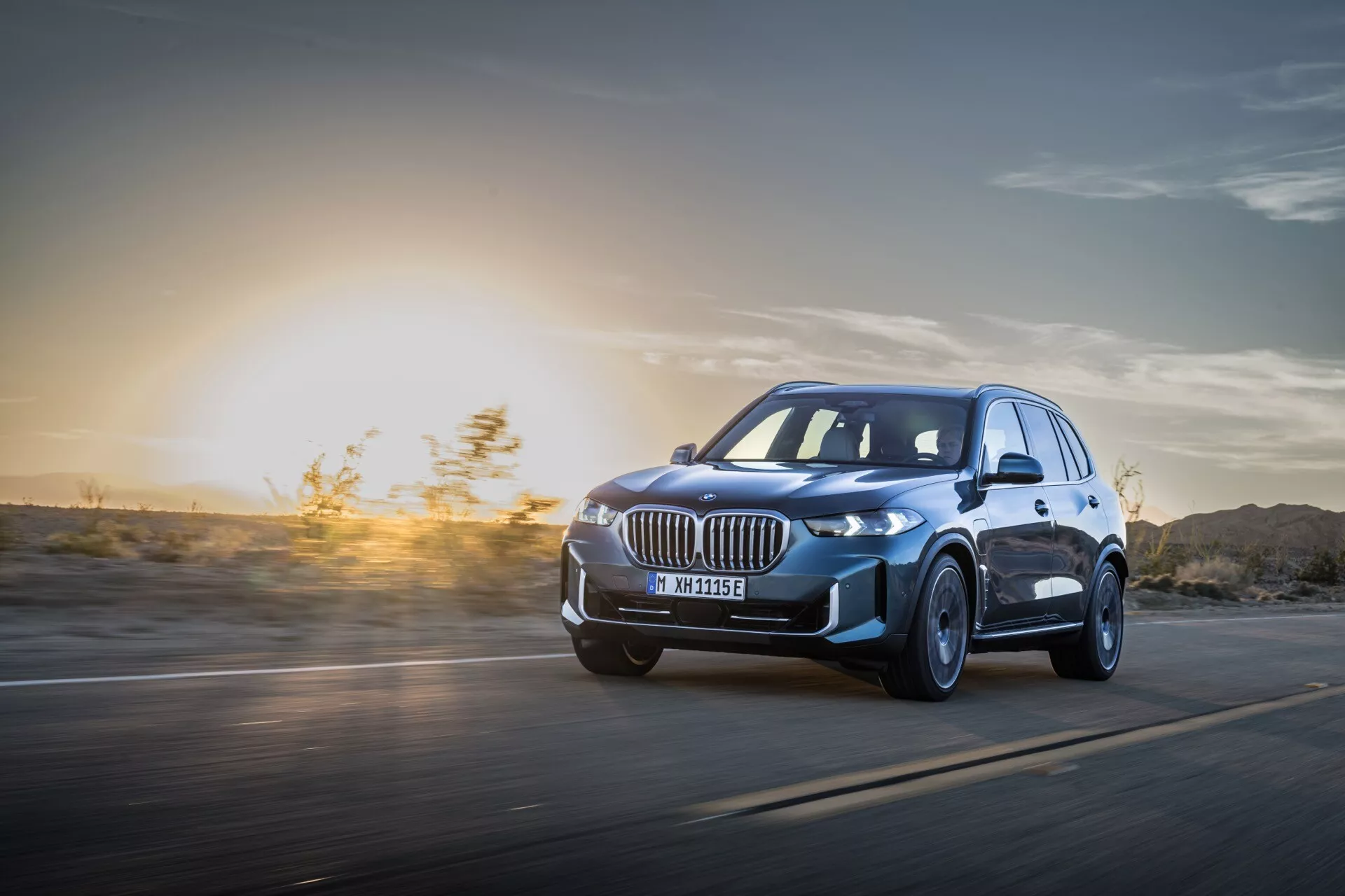 BMW X5 2023 (G05-F95 Facelift) v gibanju: Dinamičen pogled iz nizkega kota, ki poudarja LED žaromete, platišča iz zlitine in aerodinamično oblikovanje v ozadju sončnega zahoda