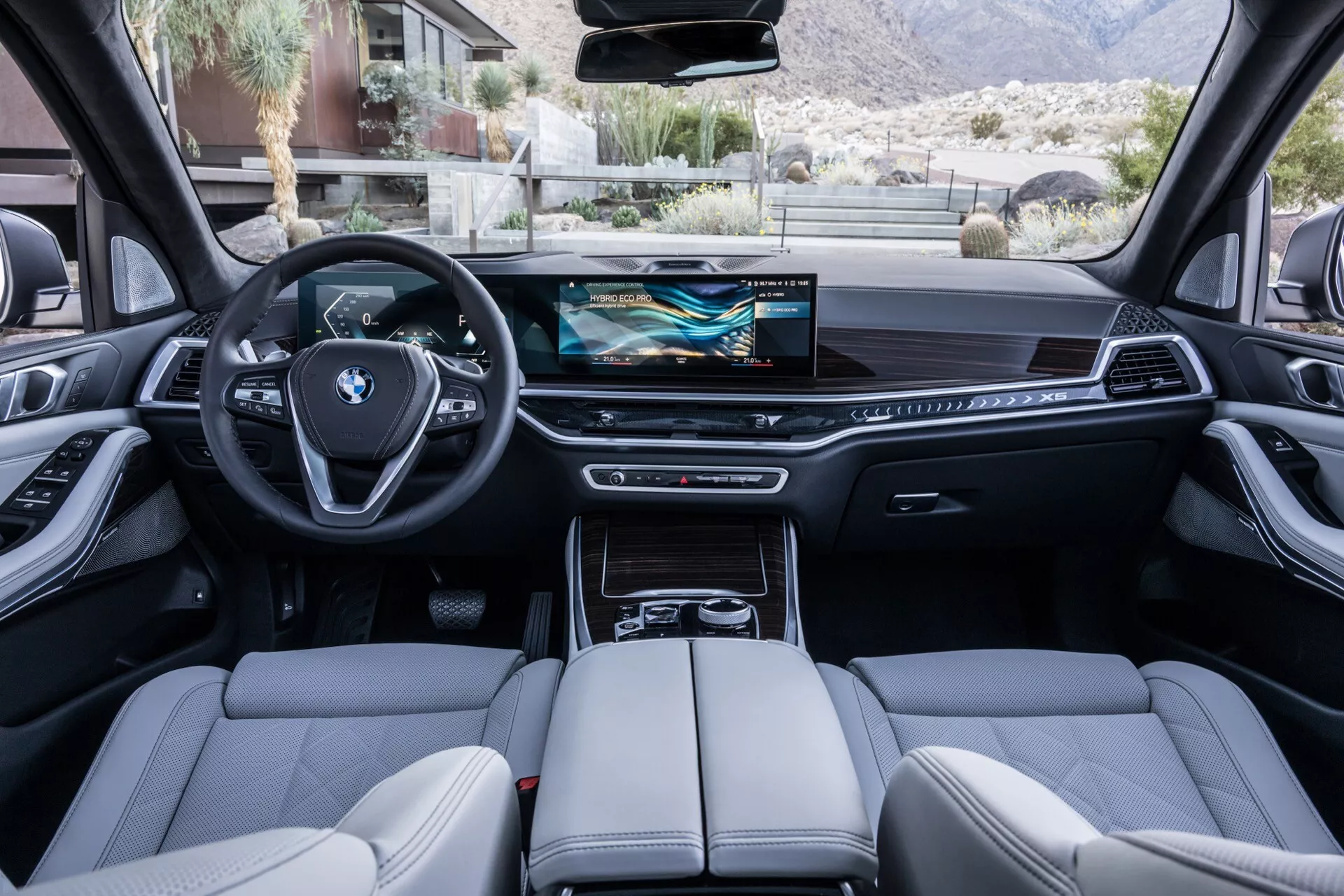Notranji pogled na BMW X5 2023 (G05-F95 Facelift) iz zadnjega sedeža, prikazuje športni volan s tremi kraki z integriranimi multifunkcijskimi gumbi, digitalni instrumentni vmesnik, širok zaslon na dotik za infotainment, minimalistične nadzorne elemente na armaturni plošči, elektronski izbirnik prestav, sedeže visoke kakovosti s podrobnimi šivi, panoramsko streho in napredne tehnološke značilnosti