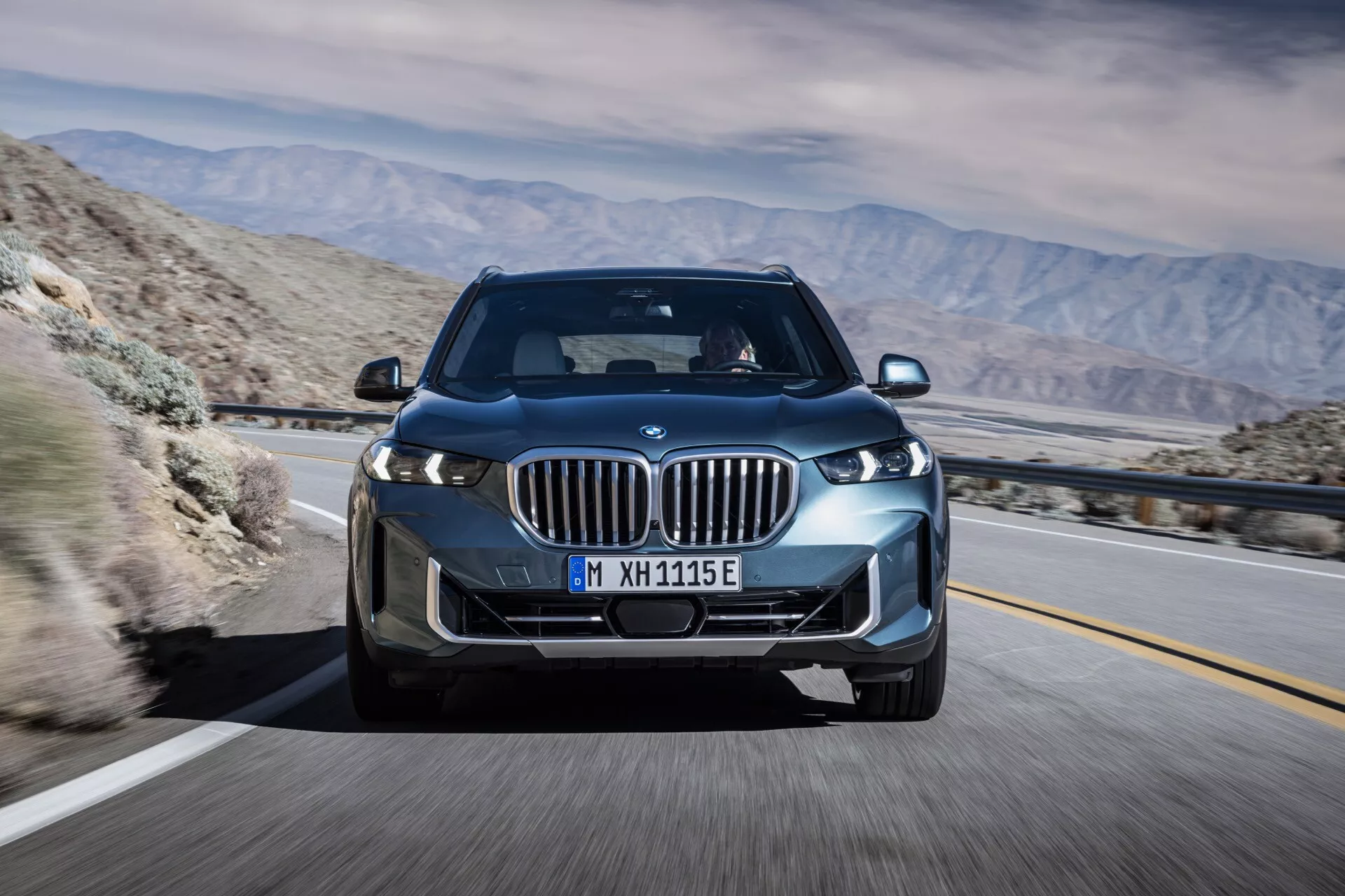 BMW X5 2023 (G05 F95 prenova) v gibanju, posnet iz kota tri četrtine spredaj z izpostavitvijo njegovih LED žarometov, ledvičke z aktivnimi lamelami, litih kolesih in elegantnim aerodinamičnim dizajnom proti gorski puščavski kulisi, kar nakazuje napredno tehnologijo in potencialne električne ali hibridne zmogljivosti