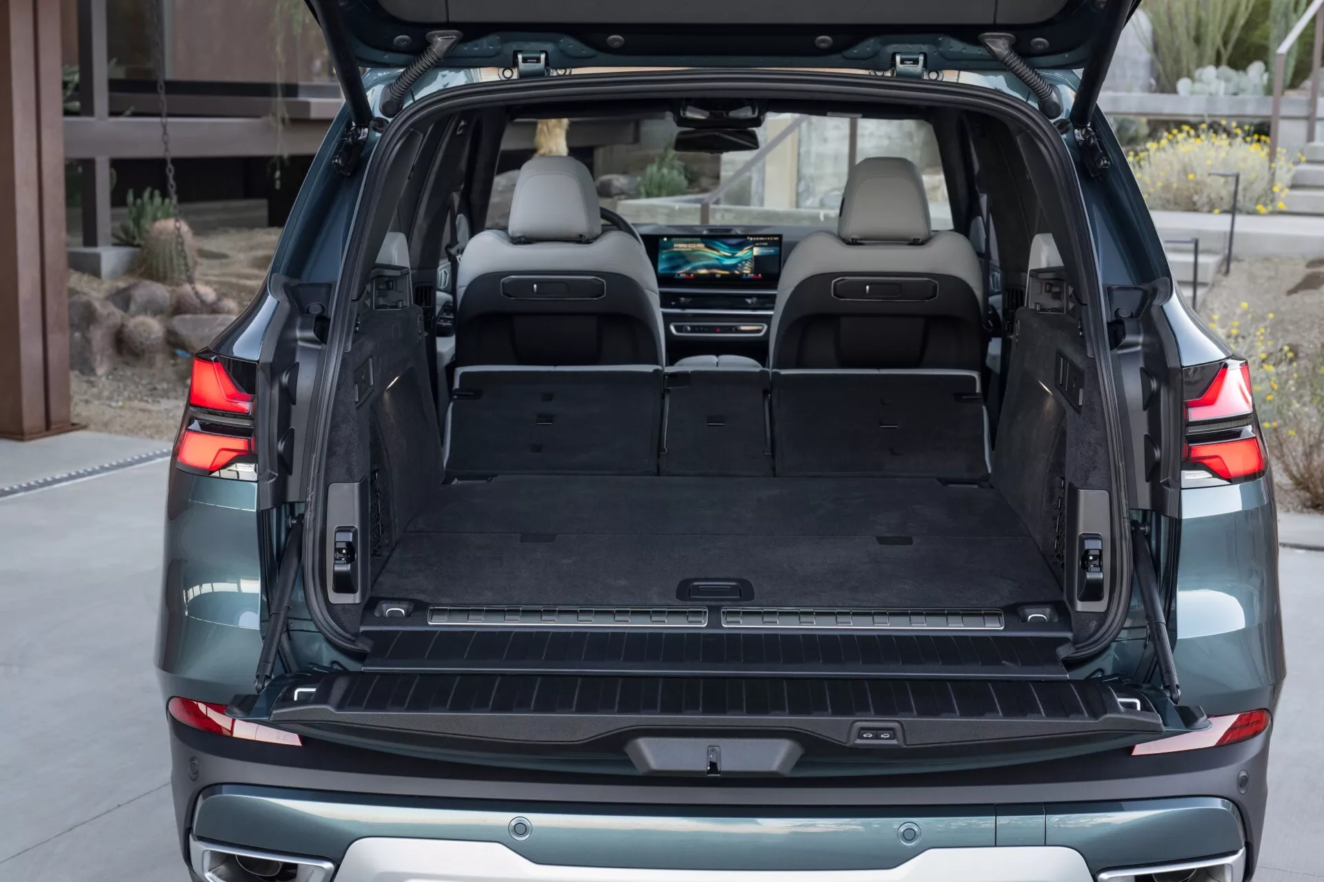 BMW X5 2023 (G05-F95 Facelift) prikazuje prostorno in dobro osvetljeno zadnjo tovorno površino s ploščatim dnom, dvignjenimi sedeži druge vrste, razkošno notranjostjo iz svetlega usnja, vidnimi točkami za pritrditev tovora in 12V vtičnico, ob robu z zaščito odbijača v stanovanjskem okolju