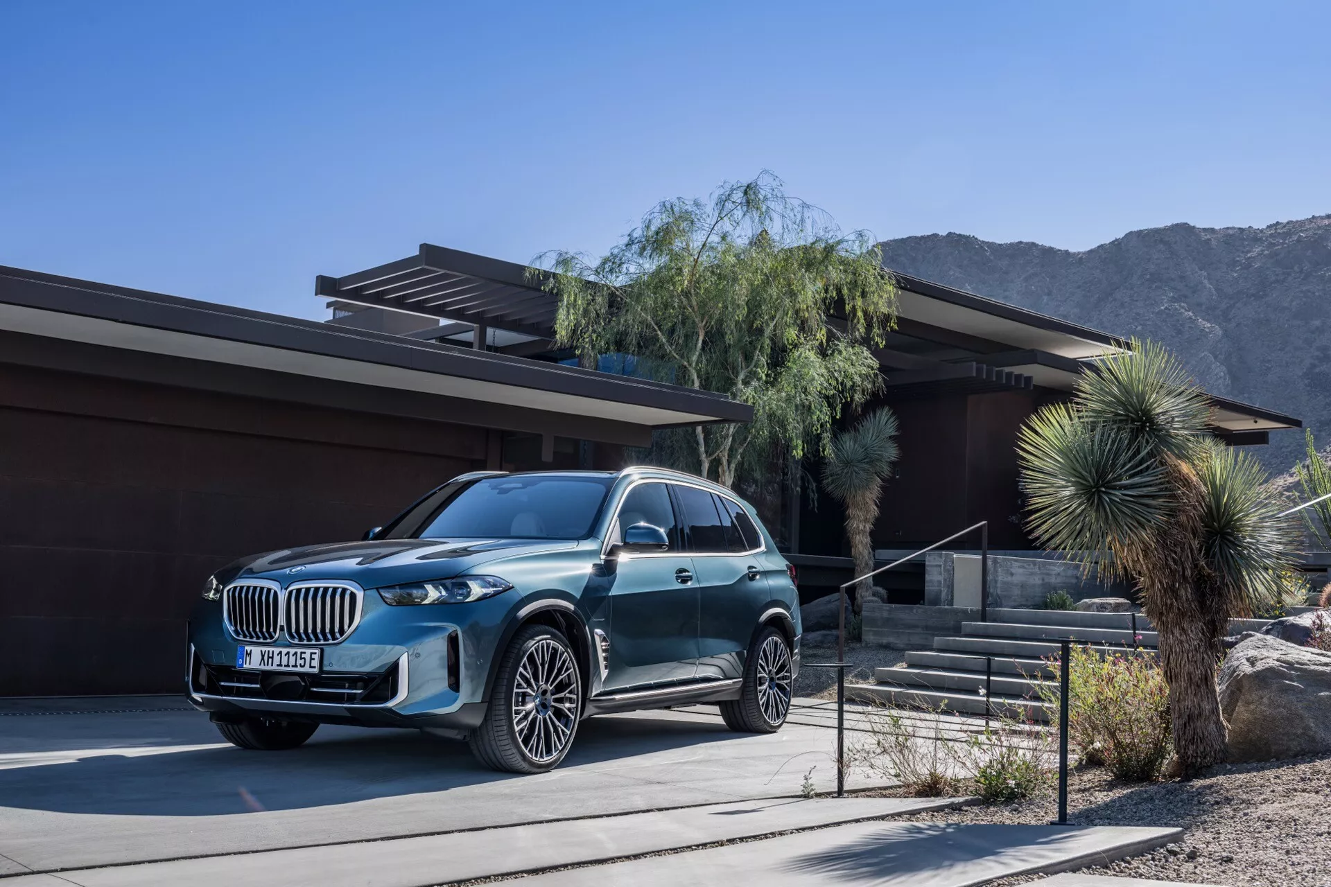 Luksuzni SUV BMW X5 2023 (G05-F95 Facelift) parkiran pred sodobno hišo z ozadjem puščavske in gorske pokrajine, z metalik zaključkom, veliko rešetko, LED žarometi in večkrakimi zlitimi kolesi