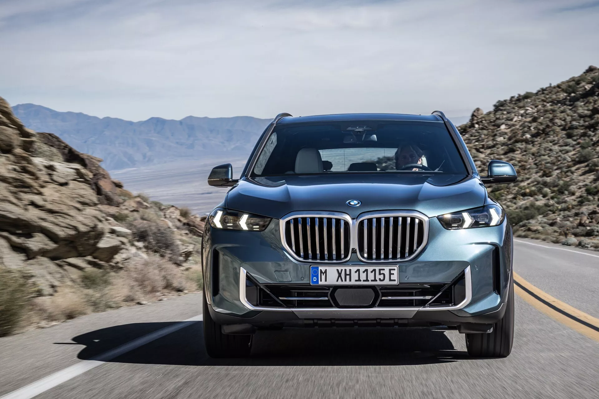 Moderno luksuzno SUV vozilo BMW X5 2023 (G05-F95-Facelift) v gibanju, s široko ledvičasto rešetko, elegantnimi LED žarometi in naprednimi tehnološkimi elementi, kot je sprednja kamera, prikazano iz kota sprednjega tričetrtinskega pogleda, ki prikazuje dinamično držo vozila in oblikovalske podrobnosti, kot so oblikovan pokrov motorja in blatniki.