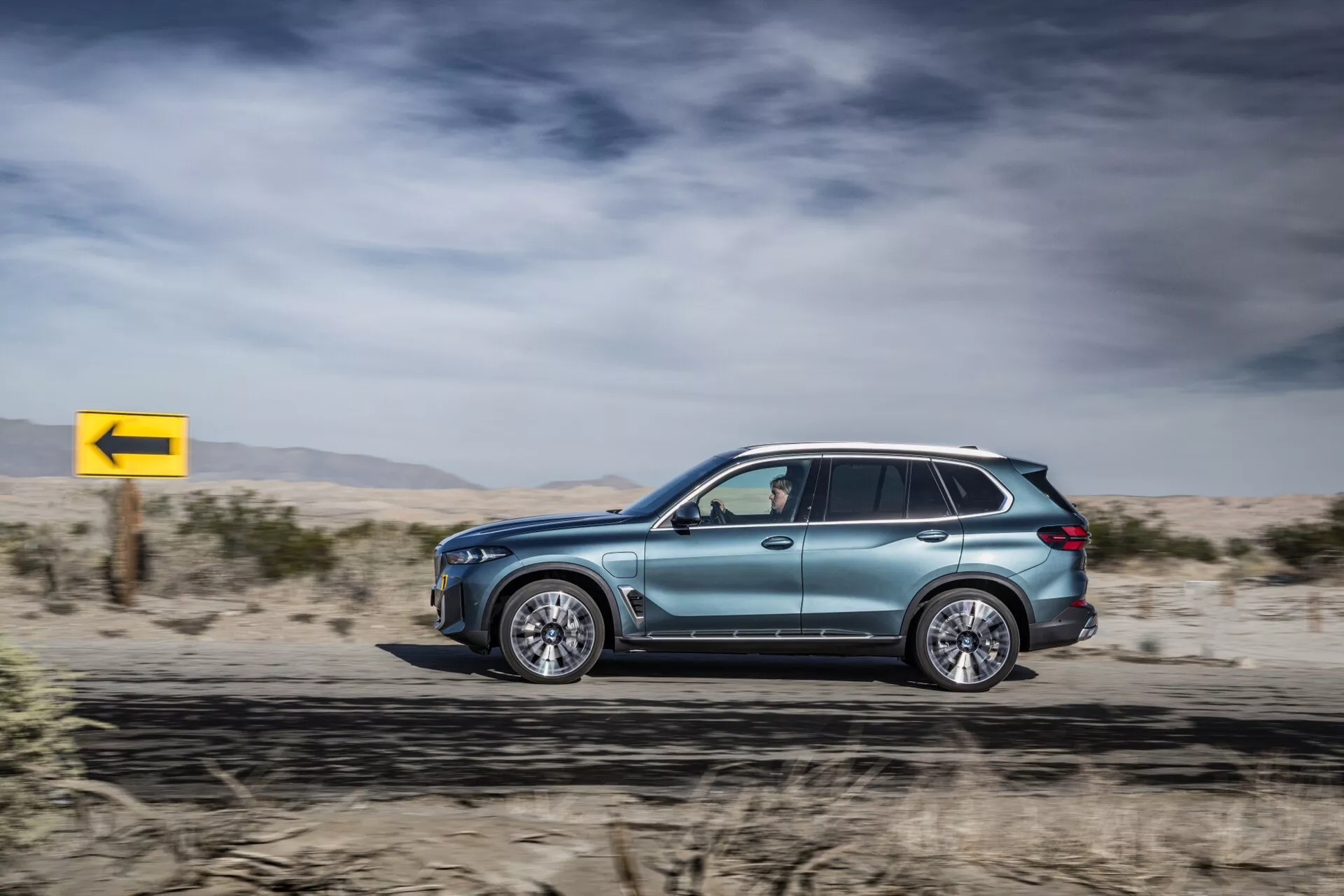 Bočni profil BMW X5 2023 (G05-F95 Facelift) v gibanju, ki prikazuje njegovo kovinsko barvo, aerodinamično obliko, luksuzna zlitinska kolesa, tonirana okna z ogledali s smerniki, strešne nosilce in napredno LED tehnologijo, proti puščavskemu ozadju z dinamičnim rumenim znakom za levi zavoj.
