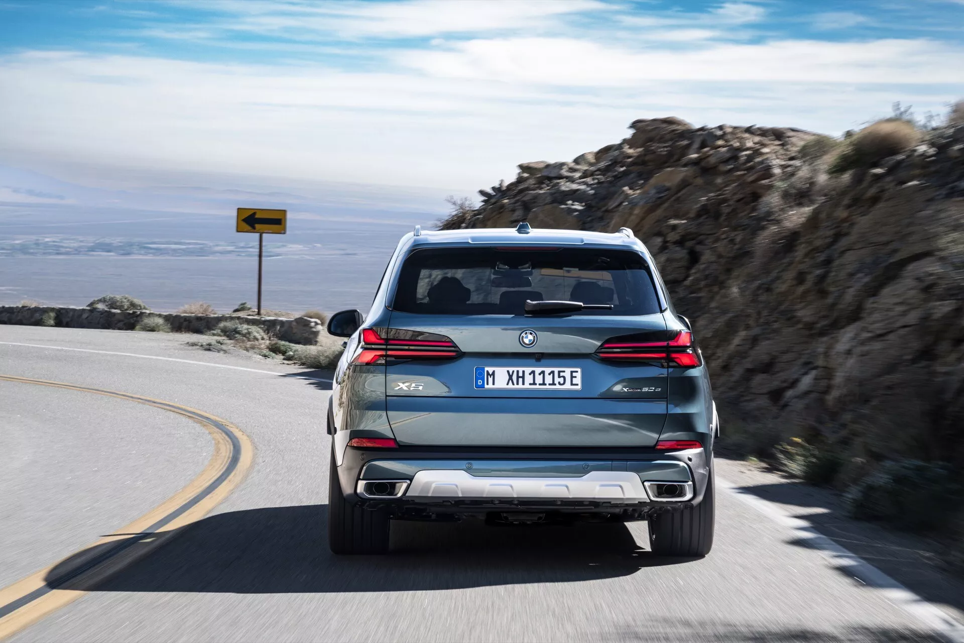Facelift BMW X5 (G05-F95) 2023 xDrive45e z M Sport paketom, ki se oddaljuje po gorski cesti, prikazuje svoj PHEV status z značkami 'eDrive' in 'M', dvojnimi izpušnimi cevmi, LED zadnjimi lučmi in evropsko registrsko tablico.